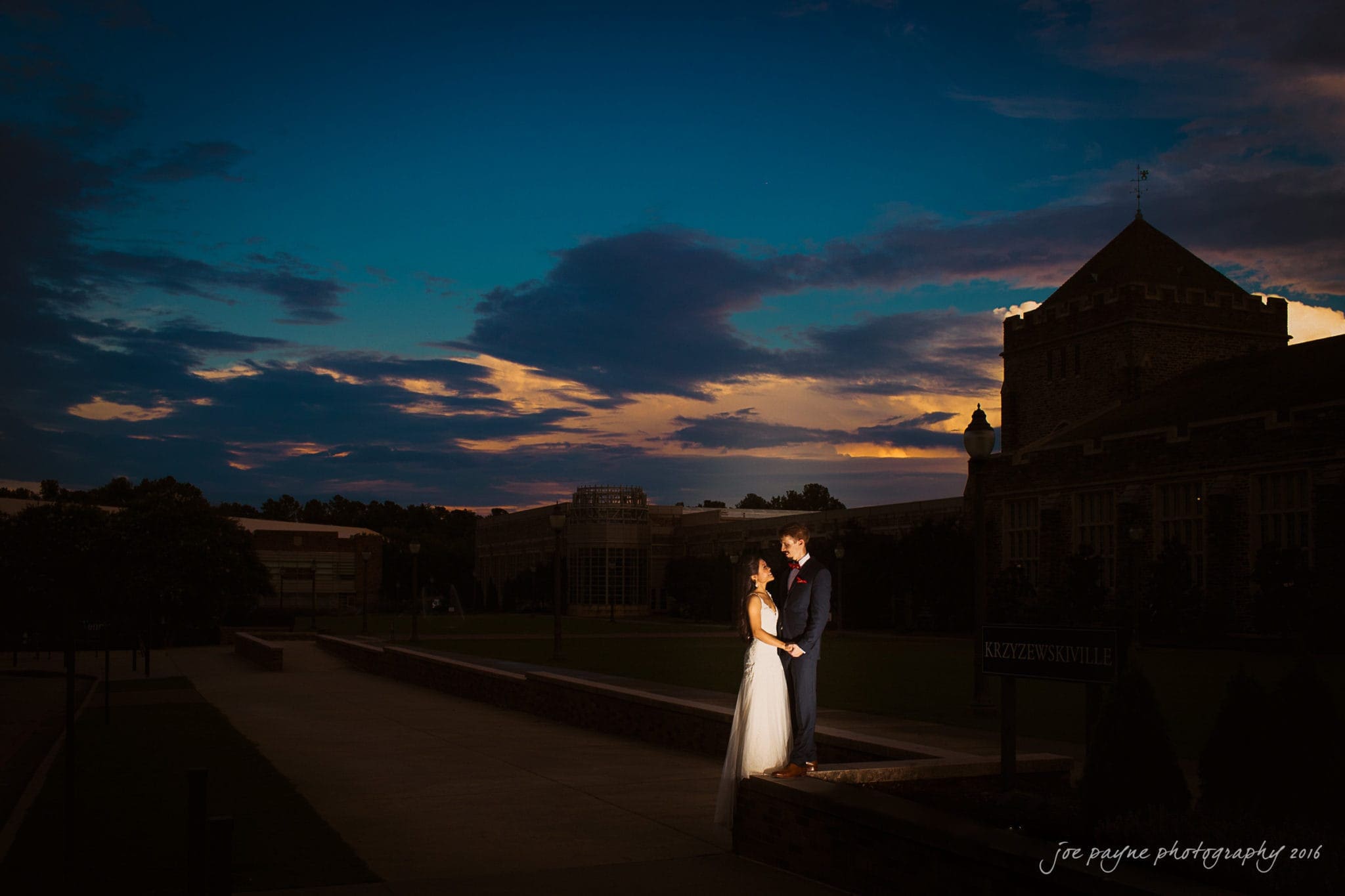 A couple stands in an outdoor courtyard, holding hands and gazing at each other at dusk. The sky is dark blue with scattered clouds and hints of orange. The silhouette of a large building is visible in the background.