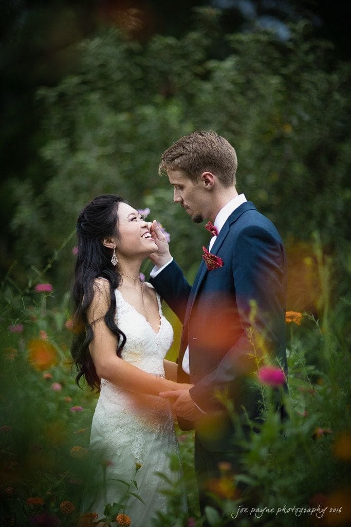 A couple stands in a lush garden, sharing an intimate moment. The woman in a white dress smiles as the man in a suit gently touches her face. Colorful flowers surround them, creating a romantic and serene setting.