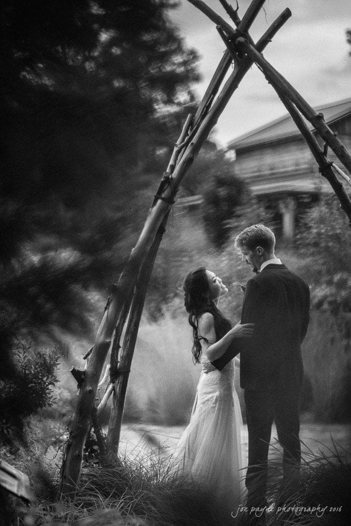 A couple in wedding attire gaze into each others eyes. They stand beneath a rustic wooden arch outdoors, surrounded by blurred natural scenery, giving the image a dreamy, romantic feel.