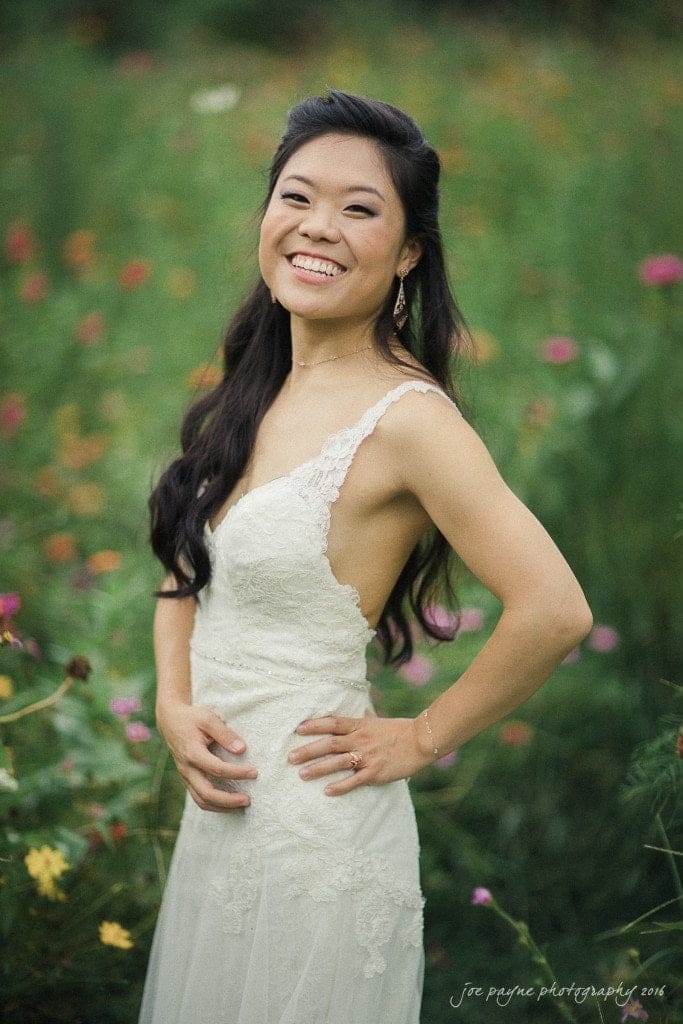 A woman in a white lace dress is smiling while standing in a garden with pink and yellow flowers in the background. Her hair is styled in long waves, and she has her hands on her hips.