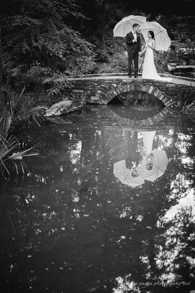 A black and white photo of a couple standing on a small stone bridge over a pond, holding umbrellas. The water reflects their image. The scene is surrounded by lush trees and foliage.