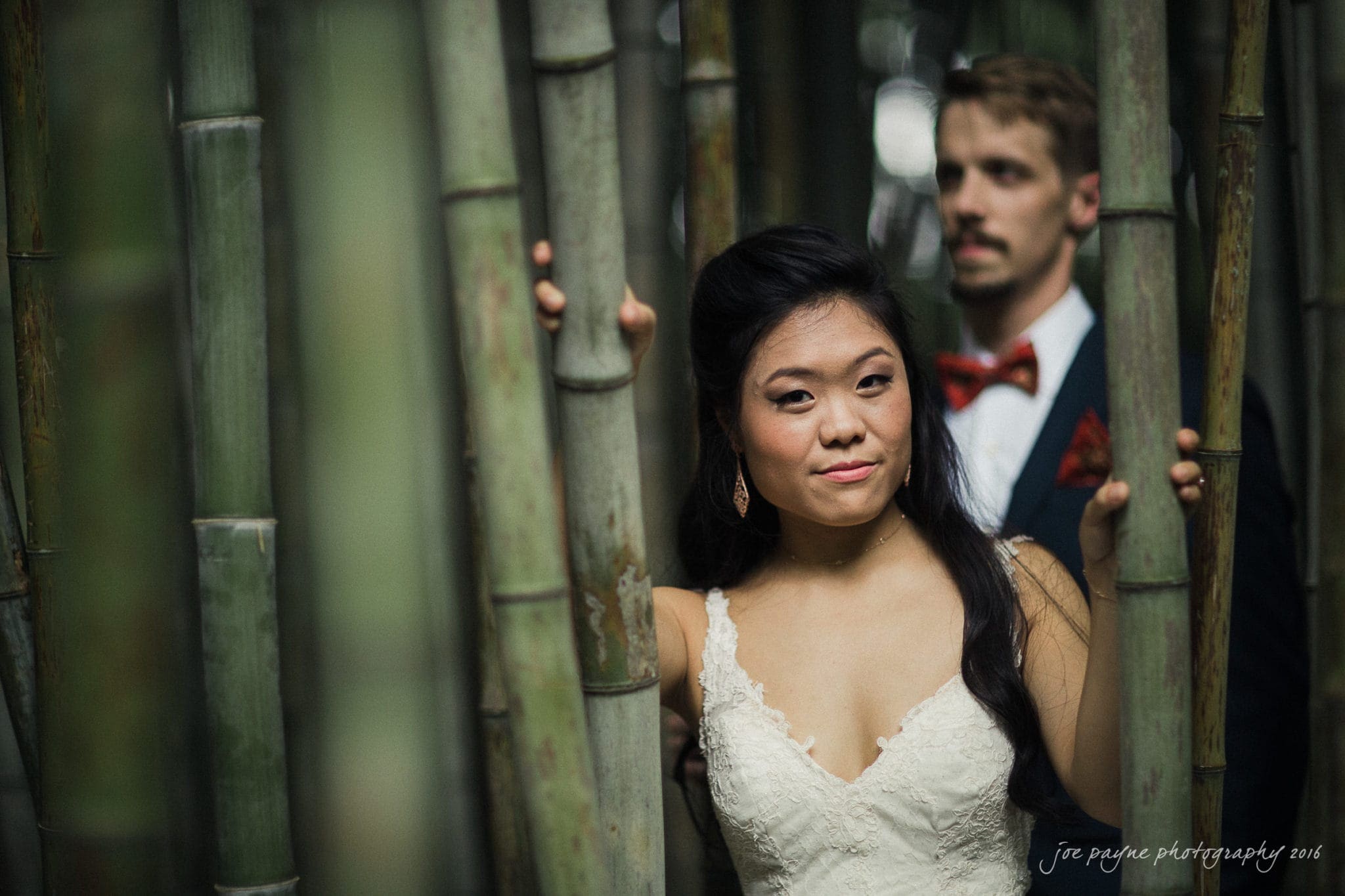 A woman in a white dress stands in a bamboo forest, looking at the camera, with a man in a suit and red bow tie behind her, partially visible among the bamboo stalks.