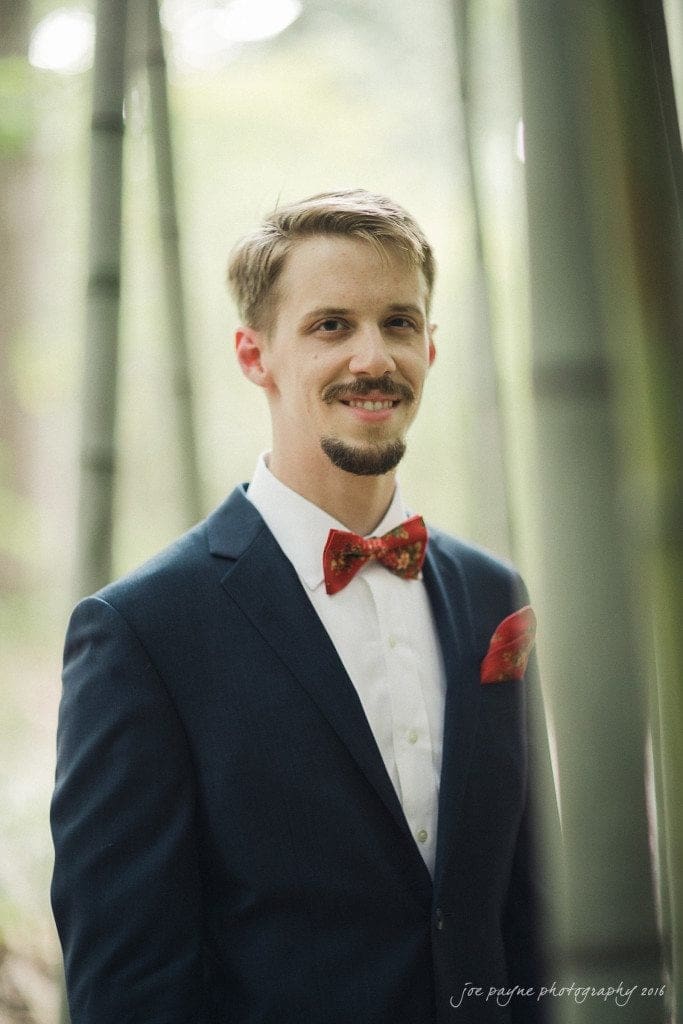 A man with short hair and a beard wears a dark suit with a white shirt and a red bow tie. He stands among tall bamboo stalks in a forest setting, looking at the camera with a slight smile.