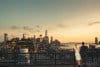 A couple stands on a rooftop, gazing at the cityscape during sunset. The skyline includes tall buildings and a river, bathed in the warm glow of early evening.