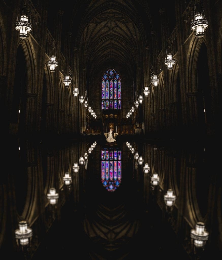 A dramatic documentary photograph of a wedding ceremony inside Duke Chapel, featuring a reflection shot where the pendant lights and altar frame the bride and groom against the large stained-glass window.