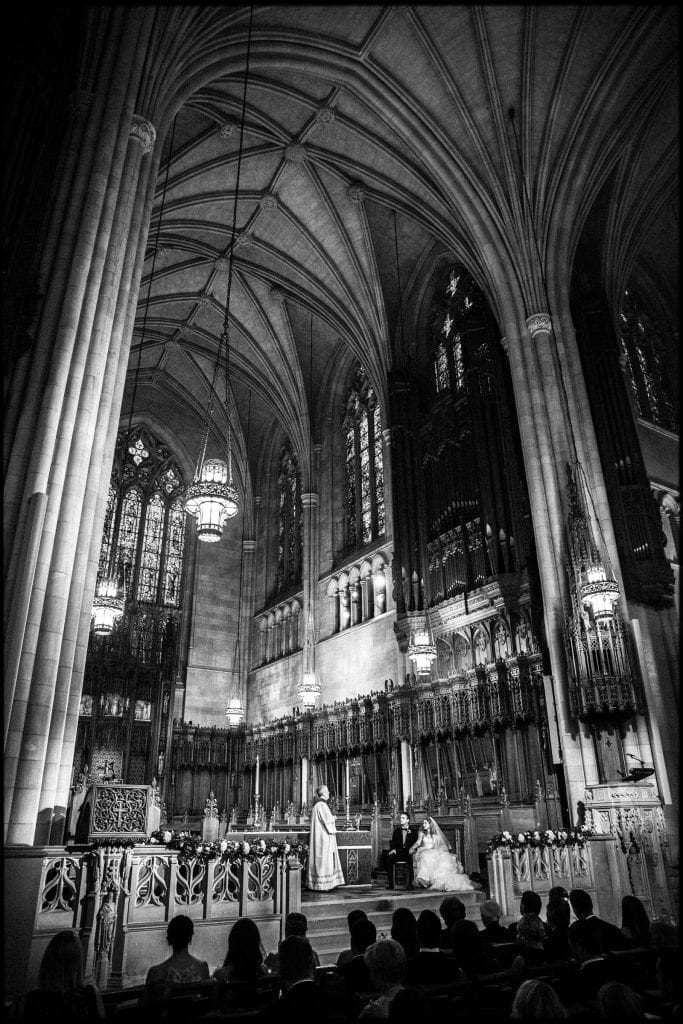 A majestic black and white vertical wedding photograph inside Duke Chapel, showing the high vaulted ceilings and carved wooden choir stalls as the bride and groom sit listening to the priest during their Catholic ceremony.