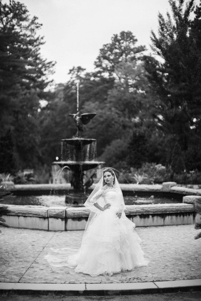 A moody black and white fine art bridal portrait of Sara posing elegantly after a rainstorm, with the historic Roney Fountain beautifully out of focus behind her at Duke University.