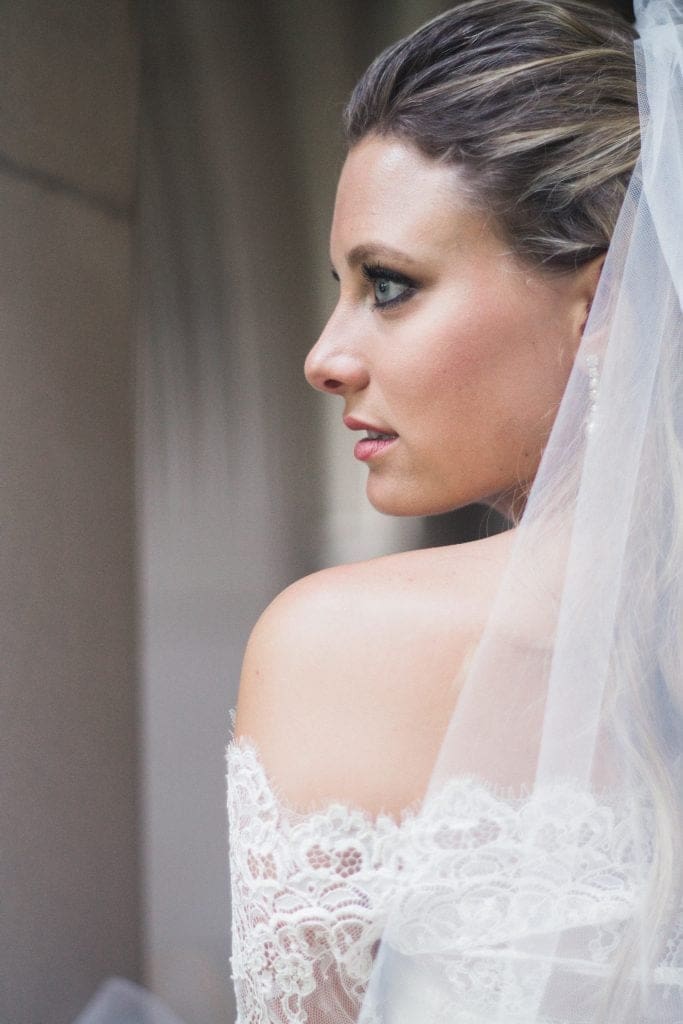A beautiful profile bridal portrait of Sara looking out over her shoulder, highlighting the elegant lace detail of her wedding dress against the historic Duke Chapel backdrop.