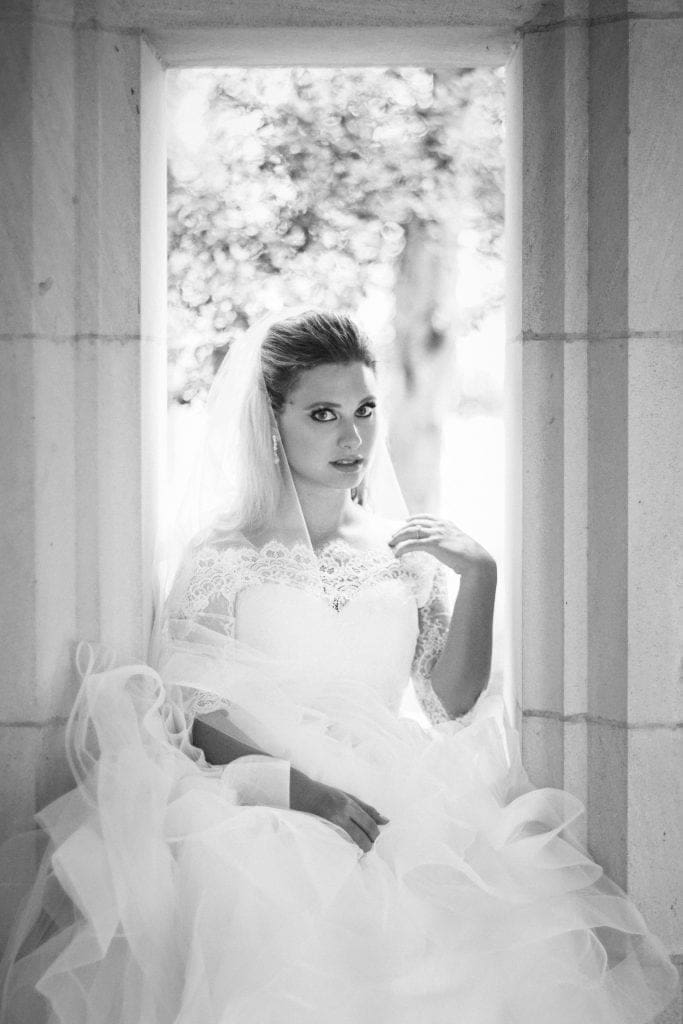 A timeless black and white fine art bridal portrait of Sara looking directly at the camera, beautifully framed by a stone window at Duke Chapel while posing with gentle, relaxed hands.