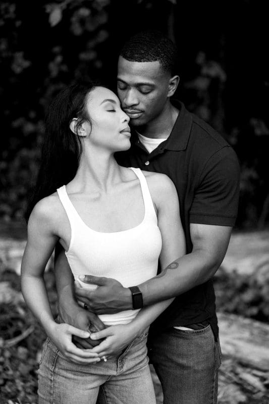 A black and white photo of a couple embracing outdoors. The woman, in a white tank top, closes her eyes while the man, in a dark polo shirt, holds her gently from behind. Trees and logs are visible in the background.