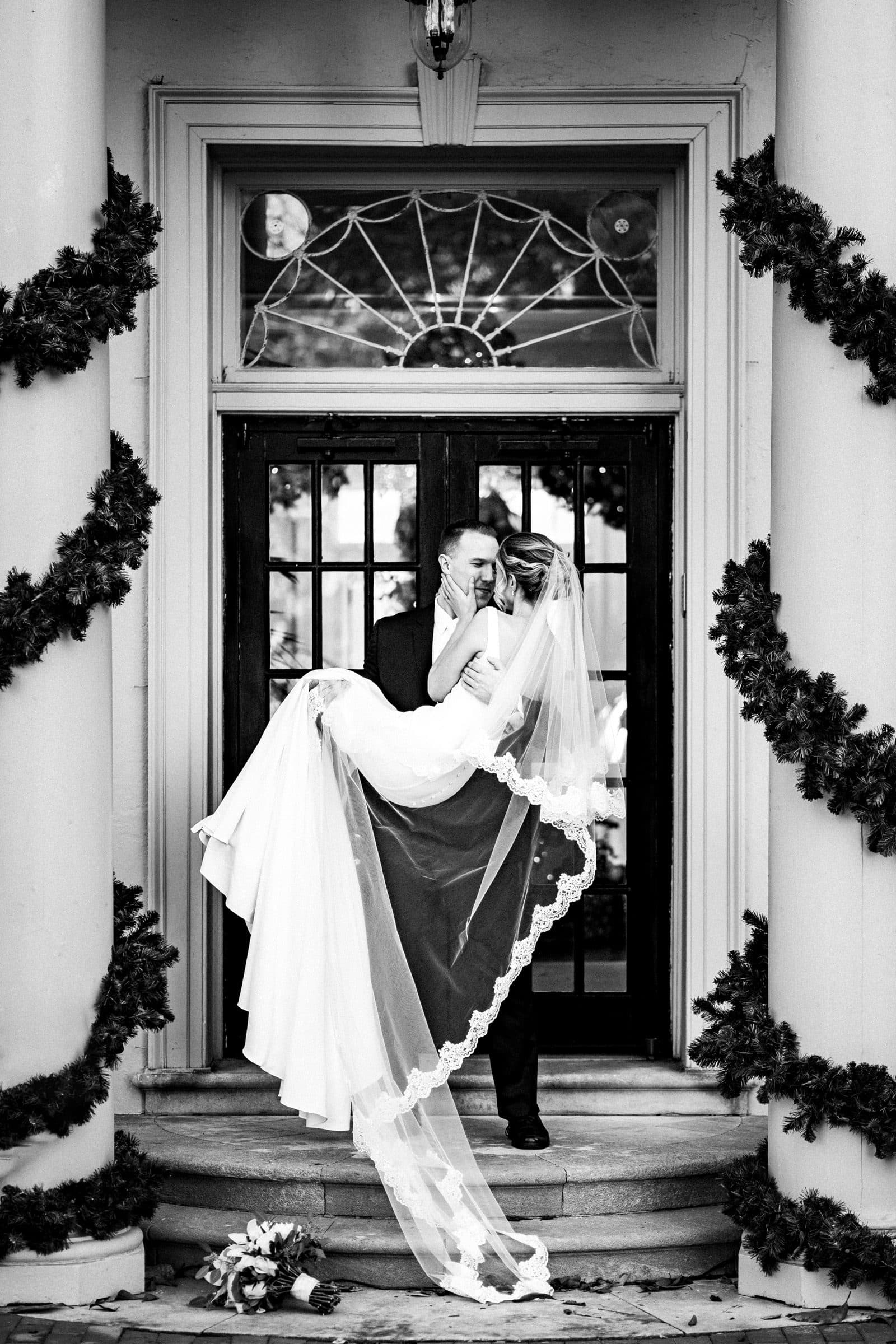 A groom lovingly carries his bride up the steps of a decorated entrance with columns and garlands. Captured by Raleigh NC wedding photographers, she wears a veil and gown, holding onto him as they share an intimate moment in this black and white wedding photograph.