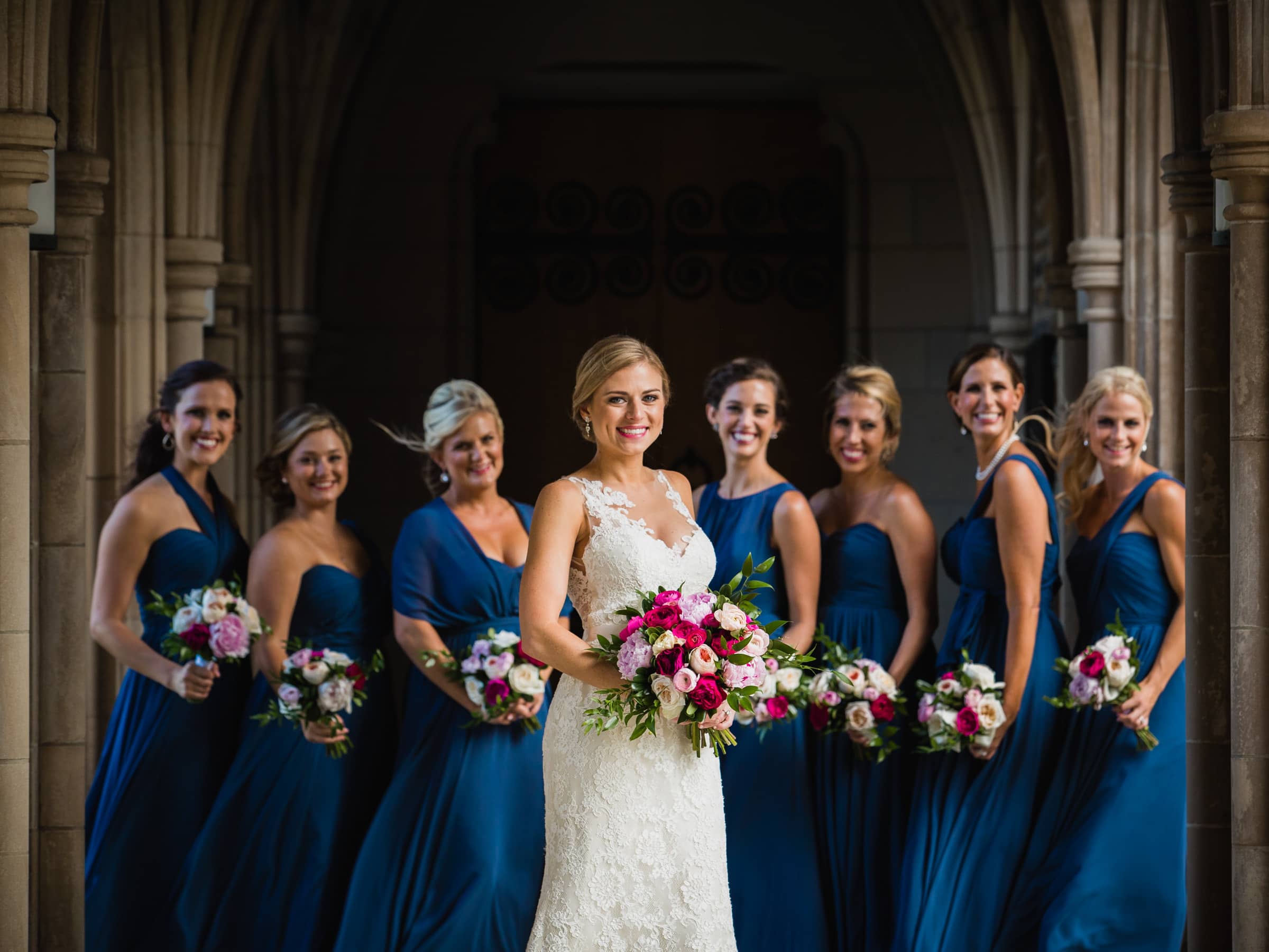 Duke Chapel Weddings A beautifully lit wedding photograph in the Duke Chapel arcades featuring a bride smiling in soft Rembrandt lighting, with her bridesmaids holding colorful bouquets in blue dresses gently out of focus behind her.