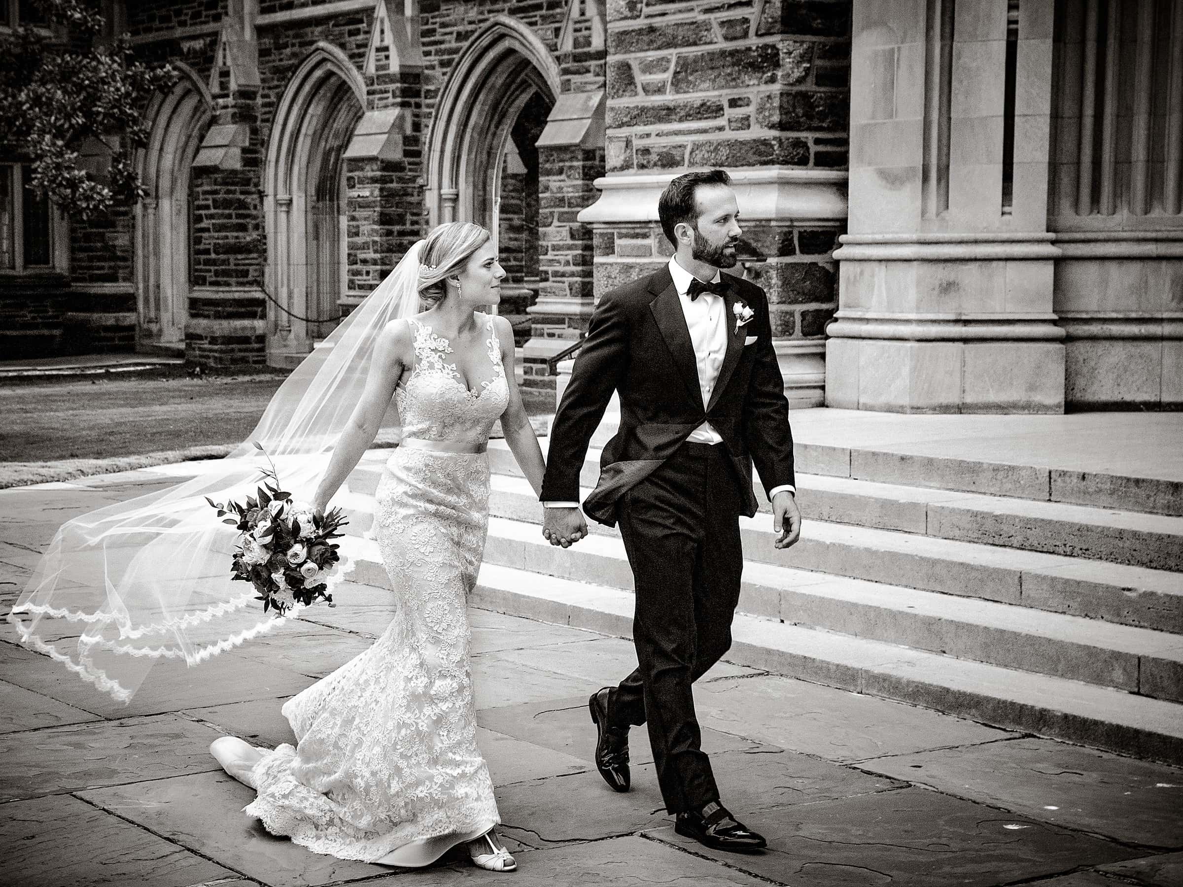 Duke Chapel Weddings A dynamic black and white candid photojournalistic wedding photograph of a couple walking briskly past the stone steps of Duke Chapel, with the bride's veil and the groom's suit jacket gently catching the wind in their wake.