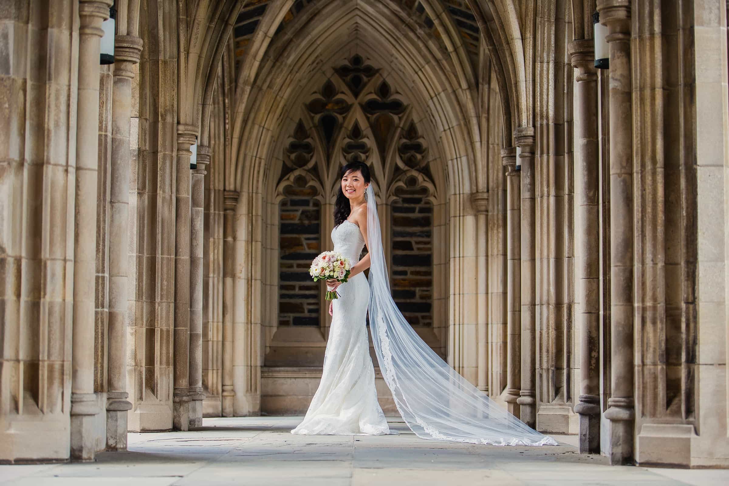 Duke Chapel Weddings A beautifully simple portrait of an Asian bride posing naturally in the Duke Chapel arcades, holding her wedding bouquet with her long veil elegantly draped behind her.