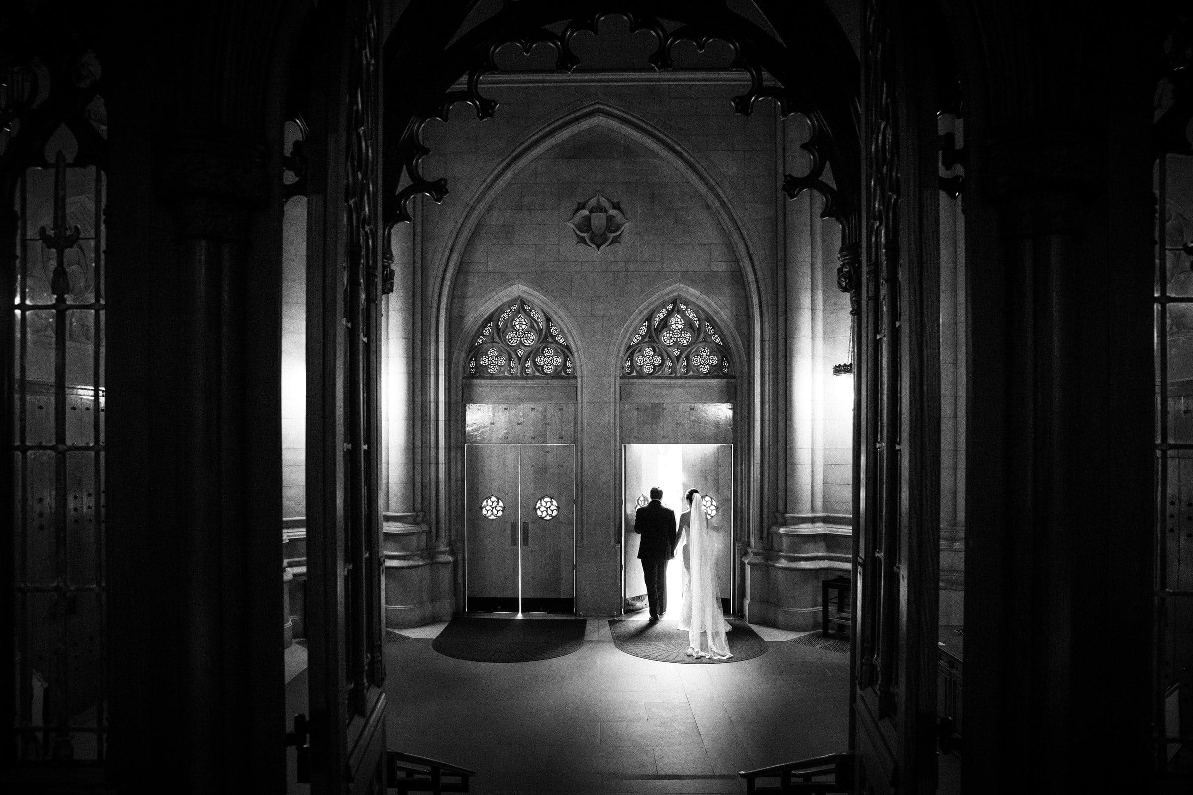 Duke Chapel Weddings A moody, wide-angle black and white wedding photograph from inside the dark Duke Chapel narthex, capturing light flooding in as the couple walks out through the grand entrance doors.