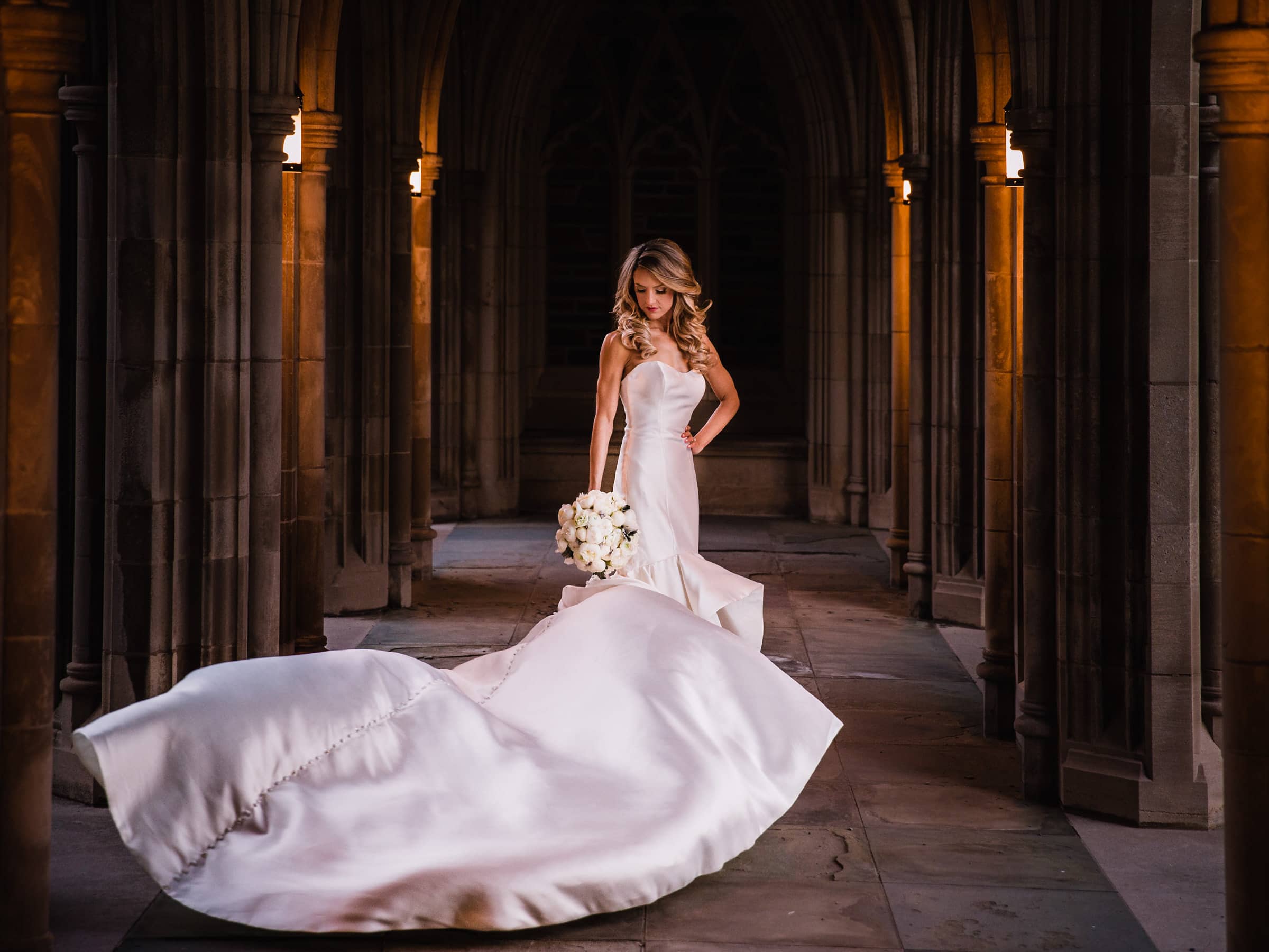 Duke Chapel Weddings A dramatic nighttime winter bridal portrait in the glowing orange lantern light of the Duke Chapel arcades, showing the bride looking down with her bouquet as a January wind catches the train of her gown before a 6pm ceremony.