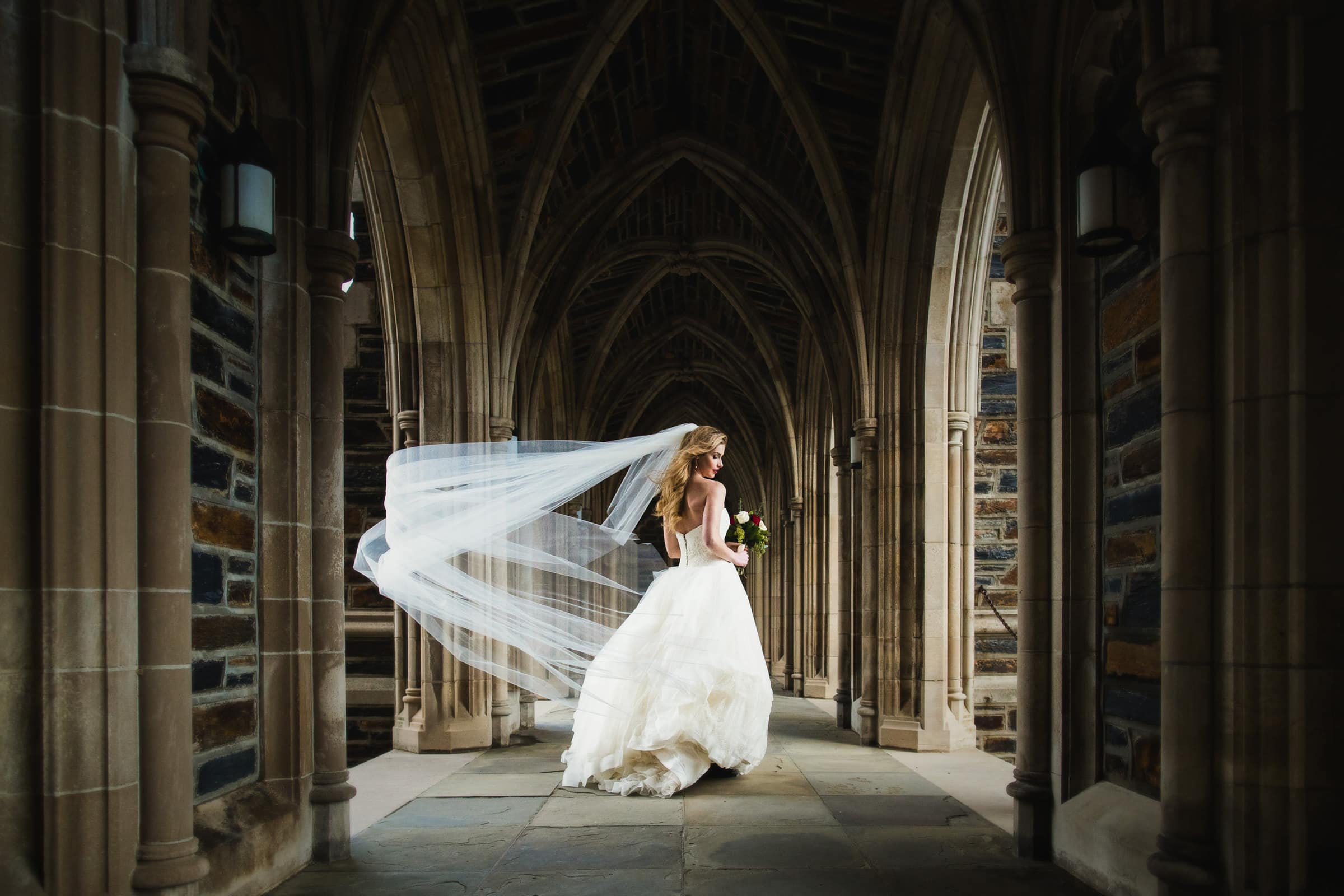 Duke Chapel Weddings A dramatic wide-angle wedding portrait in the Duke Chapel arcades, capturing the bride as a gust of wind dramatically catches her long veil beneath the historic stone arches.
