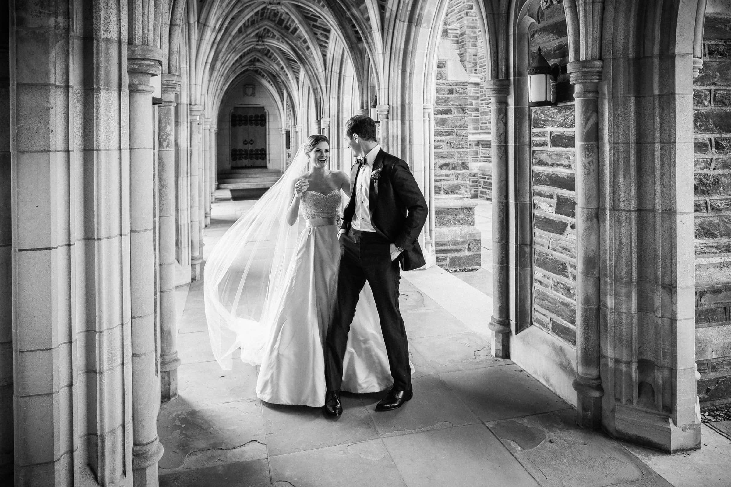 Duke Chapel Weddings A chic black and white documentary photograph of a first look in the stone arcades of Duke Chapel, taken from a three-quarter angle as the bride smiles and reaches out with her veil flowing, while the groom turns to see her in a relaxed tuxedo.