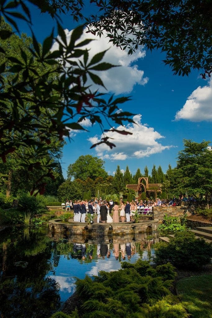 A wedding ceremony in a lush garden setting features guests gathered around a reflective pond under a bright blue sky with fluffy clouds. Captured beautifully by Raleigh NC wedding photographers, the scene is framed by green foliage, enhancing the vibrant outdoor atmosphere.