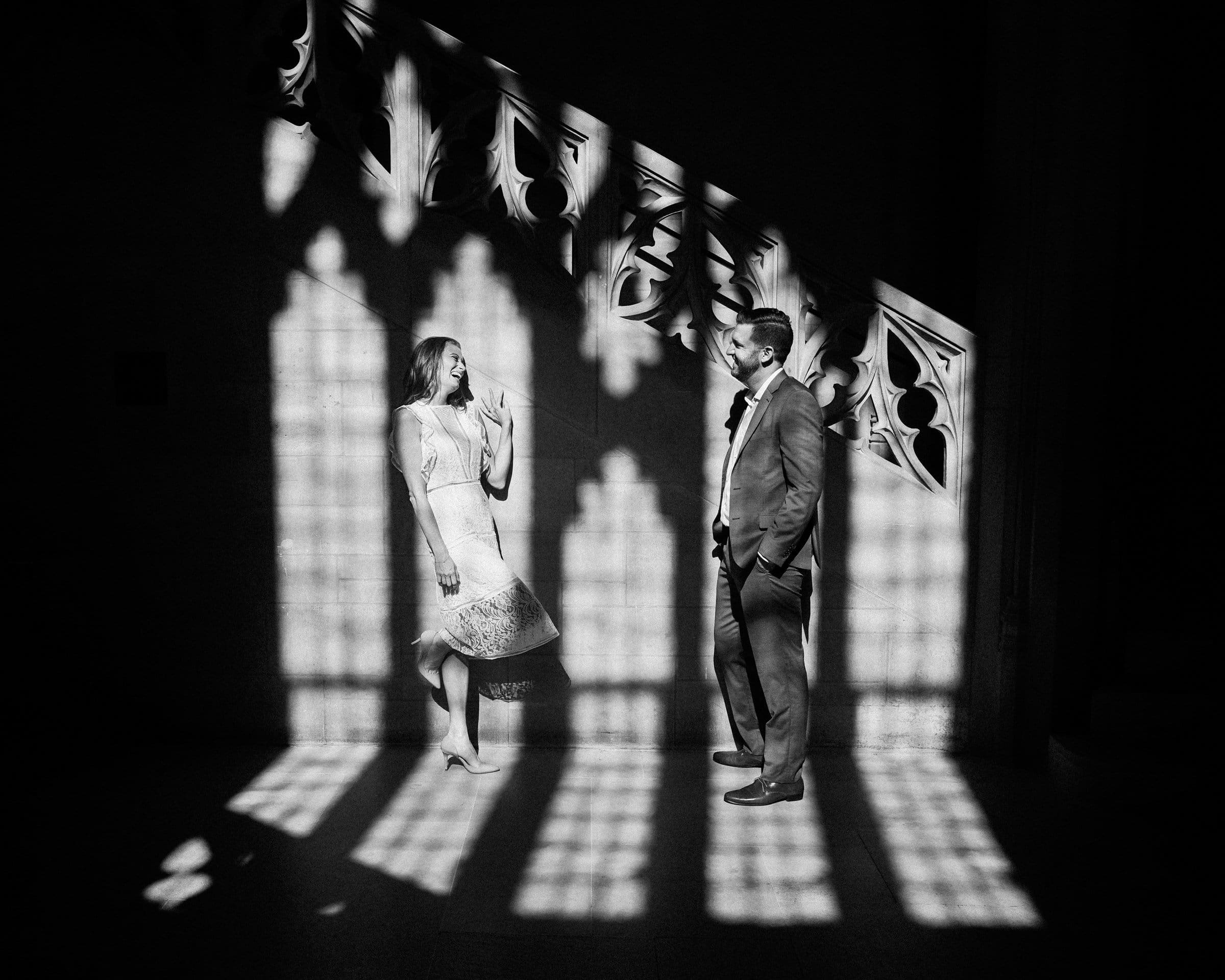 A woman in a light dress and a man in a suit stand in a dimly lit space with ornate, shadowed patterns cast across them from a window. They face each other, and the atmosphere is serene and artistic.