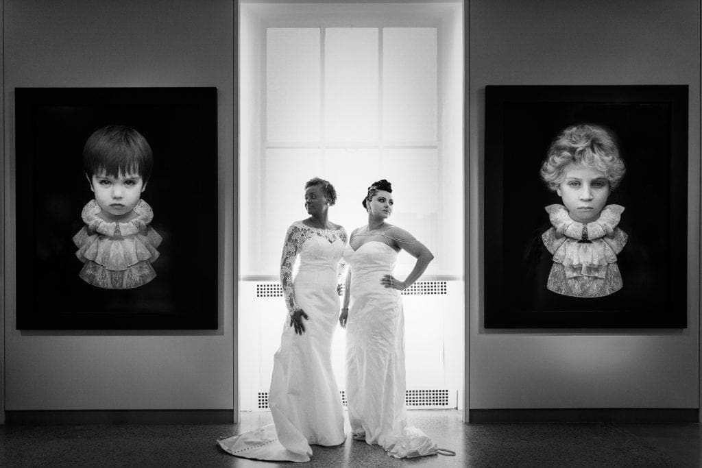 Two women in white gowns stand confidently between two large, framed portraits of solemn children, displayed in a well-lit gallery. The atmosphere is elegant and artistic.