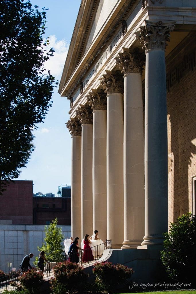 winston-salem nc wedding photography - toni & kevin A group of people in formal attire walks up the steps of a historic church with large columns and the words First Baptist Church on the facade. Trees and buildings are visible in the background.