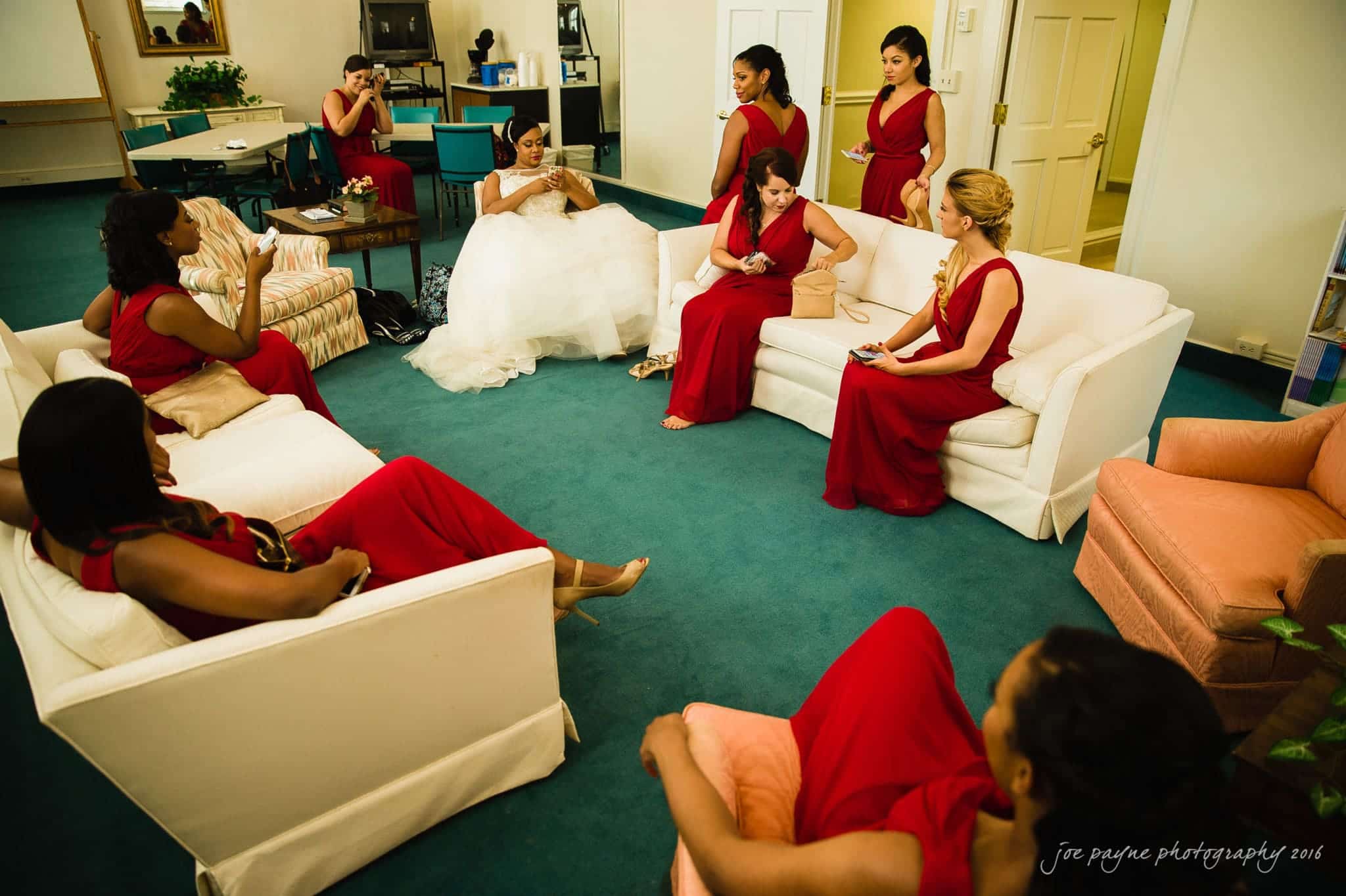 winston-salem nc wedding photography - toni & kevin A bride in a white gown sits on a sofa in a room with bridesmaids dressed in red gowns. They are gathered, chatting and preparing, with makeup and mirrors in hand. The room has teal carpet, beige sofas, and a mix of furniture.