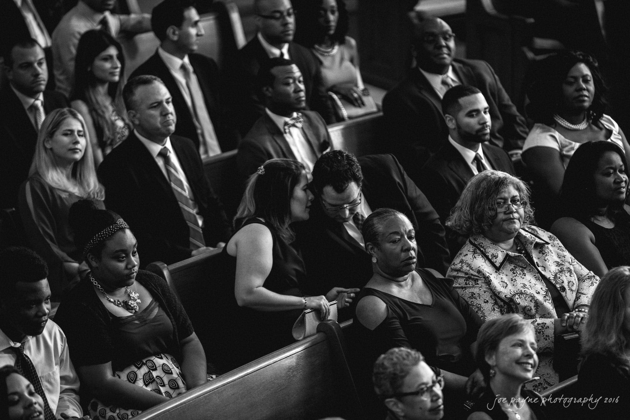 winston-salem nc wedding photography - toni & kevin A black and white photo of diverse people seated in church pews, dressed formally. Two people in the middle are leaning in and whispering to each other, creating a sense of intrigue among the attentive crowd.