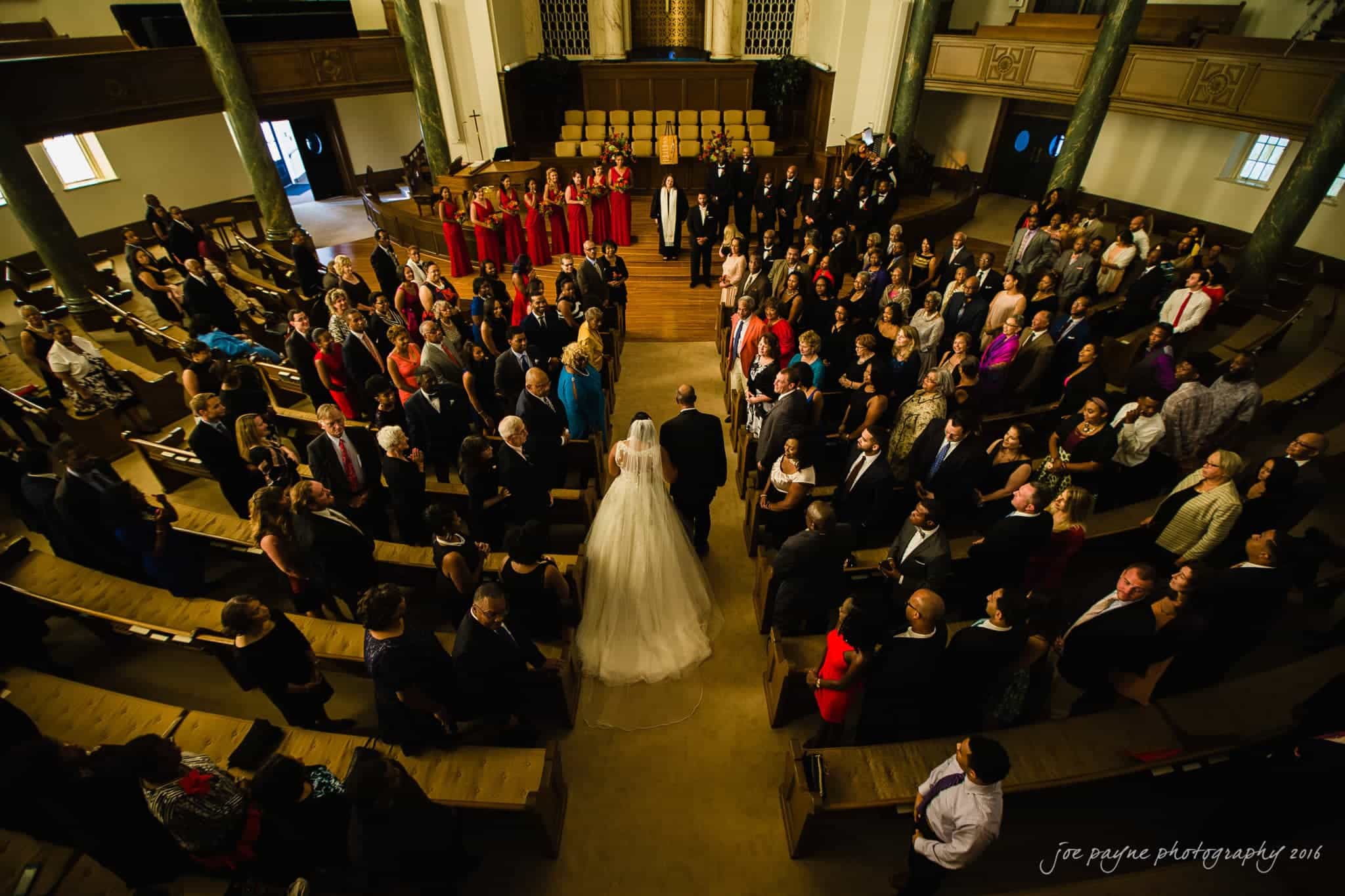 winston-salem nc wedding photography - toni & kevin A bride and groom walk down the aisle of a large, ornate church. They are surrounded by guests seated and standing, some wearing formal attire. The church features wooden pews and tall columns, with a warm, softly lit atmosphere.