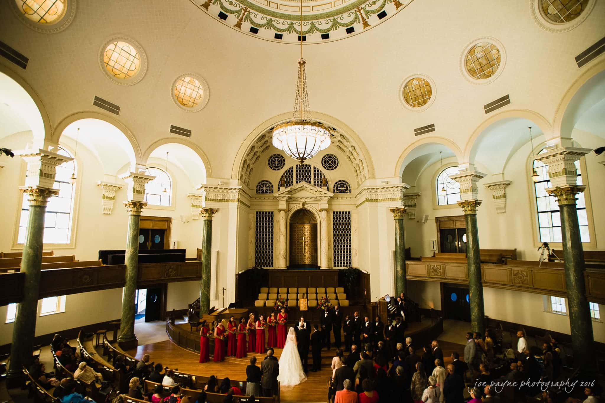 winston-salem nc wedding photography - toni & kevin A large wedding ceremony set in an ornate, circular hall with a domed ceiling and chandelier. The bridal party stands on a wooden stage, surrounded by rows of seated guests. The architecture features arches, columns, and intricate details.