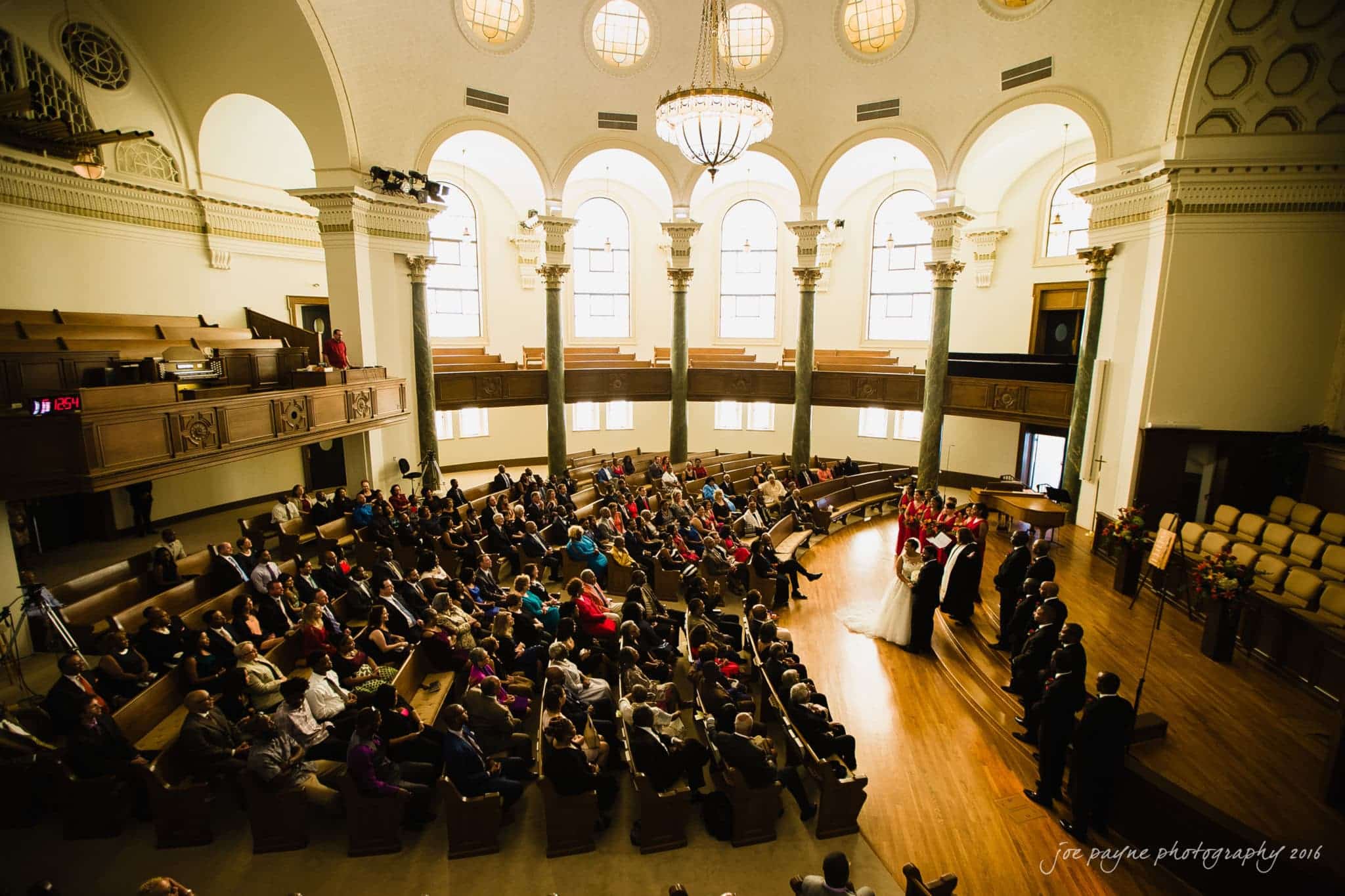 winston-salem nc wedding photography - toni & kevin A wedding ceremony in a large, elegant venue with a high ceiling, arched windows, and columns. Guests are seated in wooden pews, while the wedding party stands at the front. Natural light fills the room, enhancing the formal atmosphere.