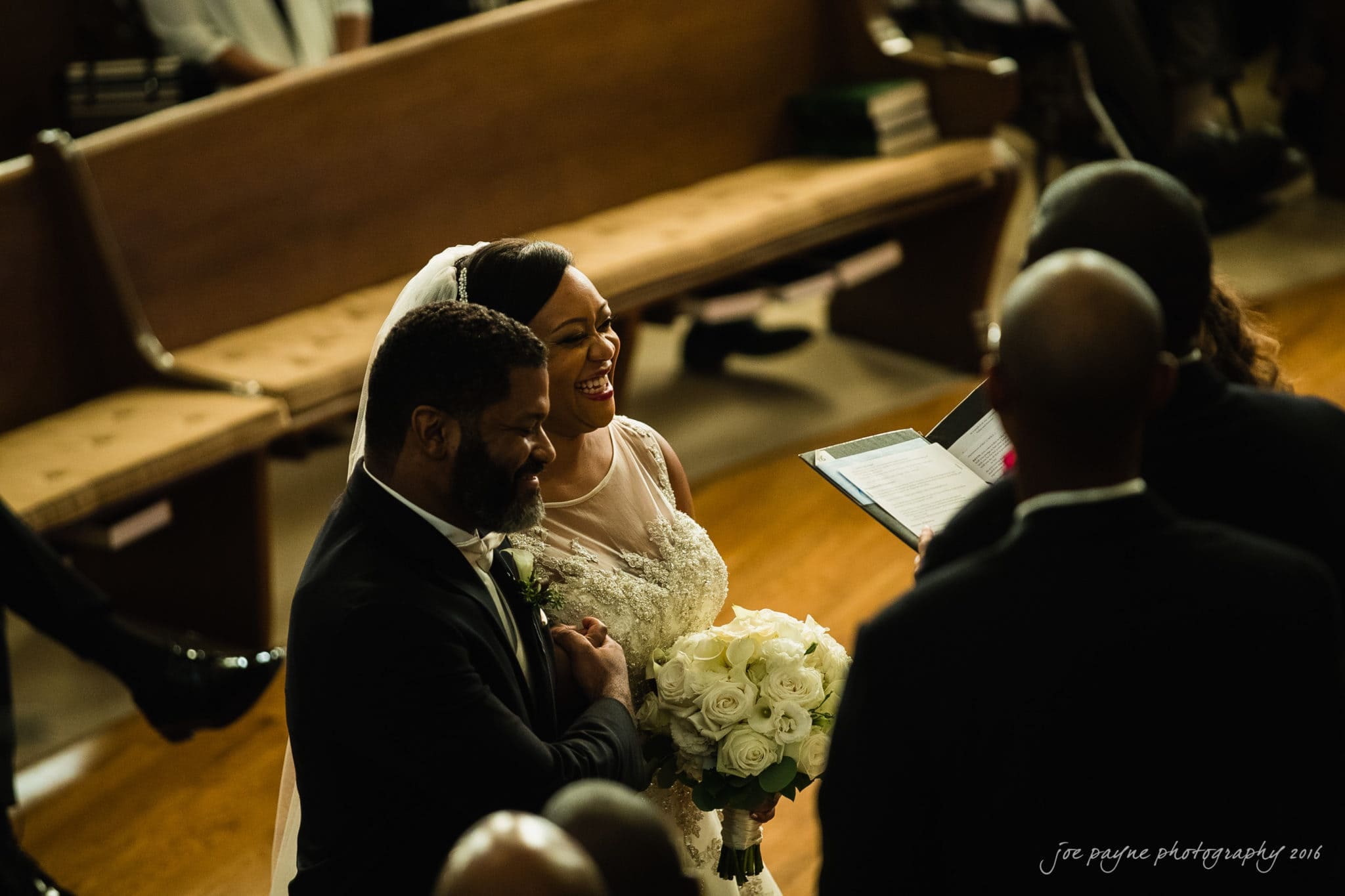 winston-salem nc wedding photography - toni & kevin A bride in a white gown and veil holds a bouquet of white flowers, walking down the aisle with a man in a black suit. They are smiling as they approach a person holding a book, possibly the officiant, during the ceremony in a dimly lit venue.