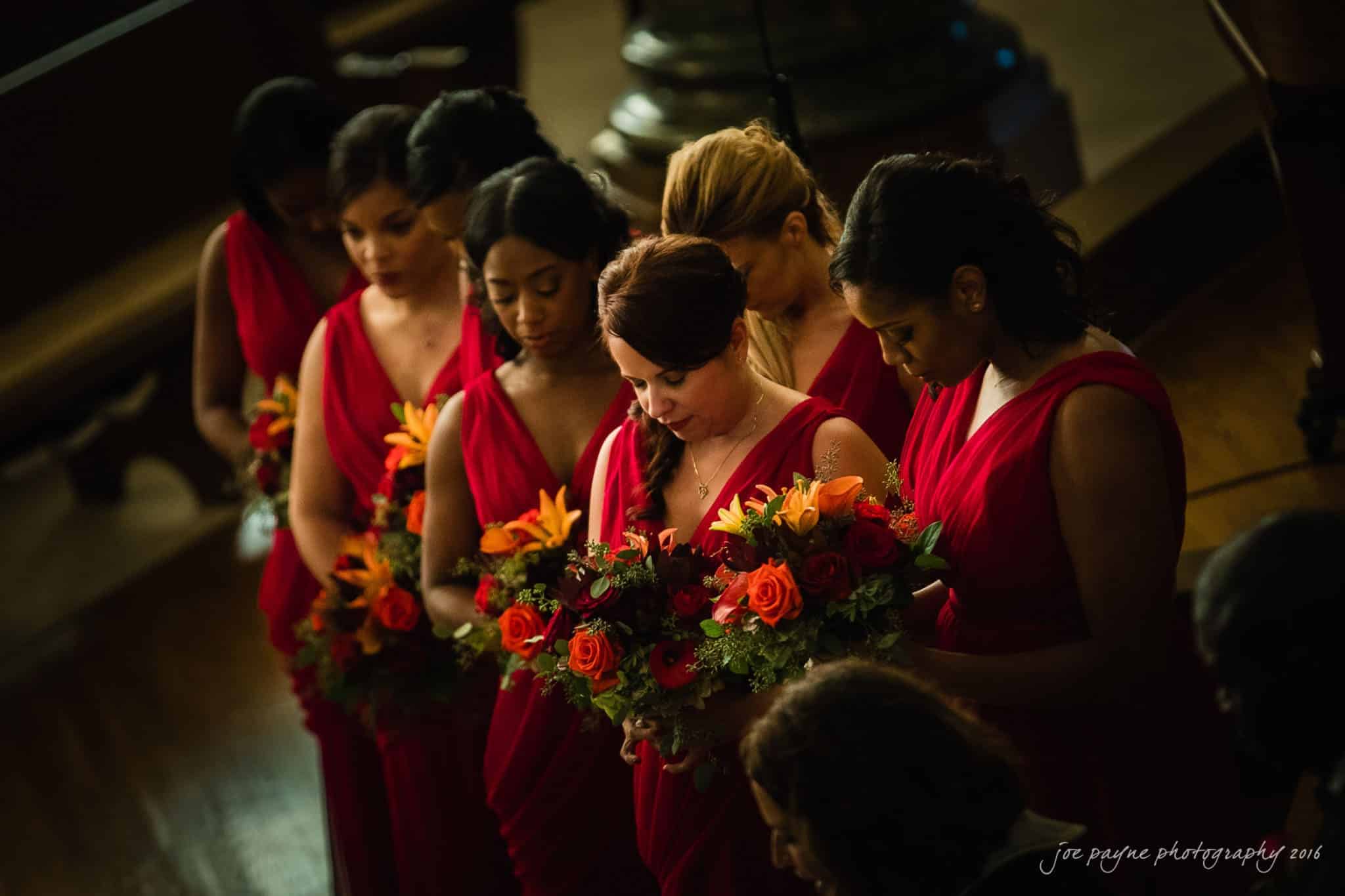 winston-salem nc wedding photography - toni & kevin A group of bridesmaids in red dresses stand closely together, holding colorful floral bouquets with orange and red flowers. They appear to be in a dimly lit indoor setting, possibly a ceremony.