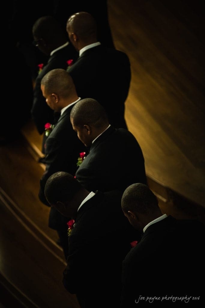 winston-salem nc wedding photography - toni & kevin A group of five people in dark suits stand in a line, holding red roses. They are positioned diagonally, facing downward, with a wooden floor in the background. The lighting casts an atmospheric shadow.