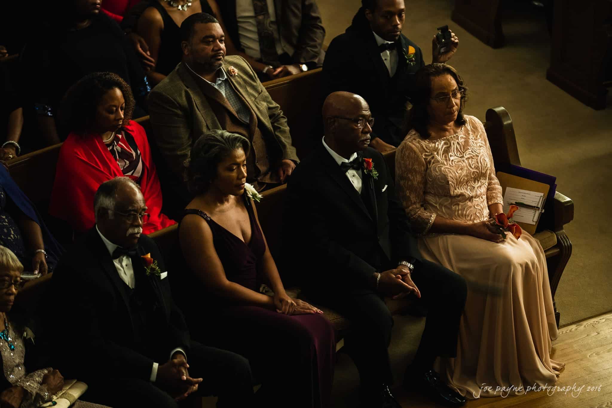winston-salem nc wedding photography - toni & kevin A group of well-dressed people sit in wooden pews at a formal event, possibly a wedding or ceremony, with attentive expressions. Men are in suits, and women wear elegant dresses. The setting is dimly lit, highlighting the attendees.