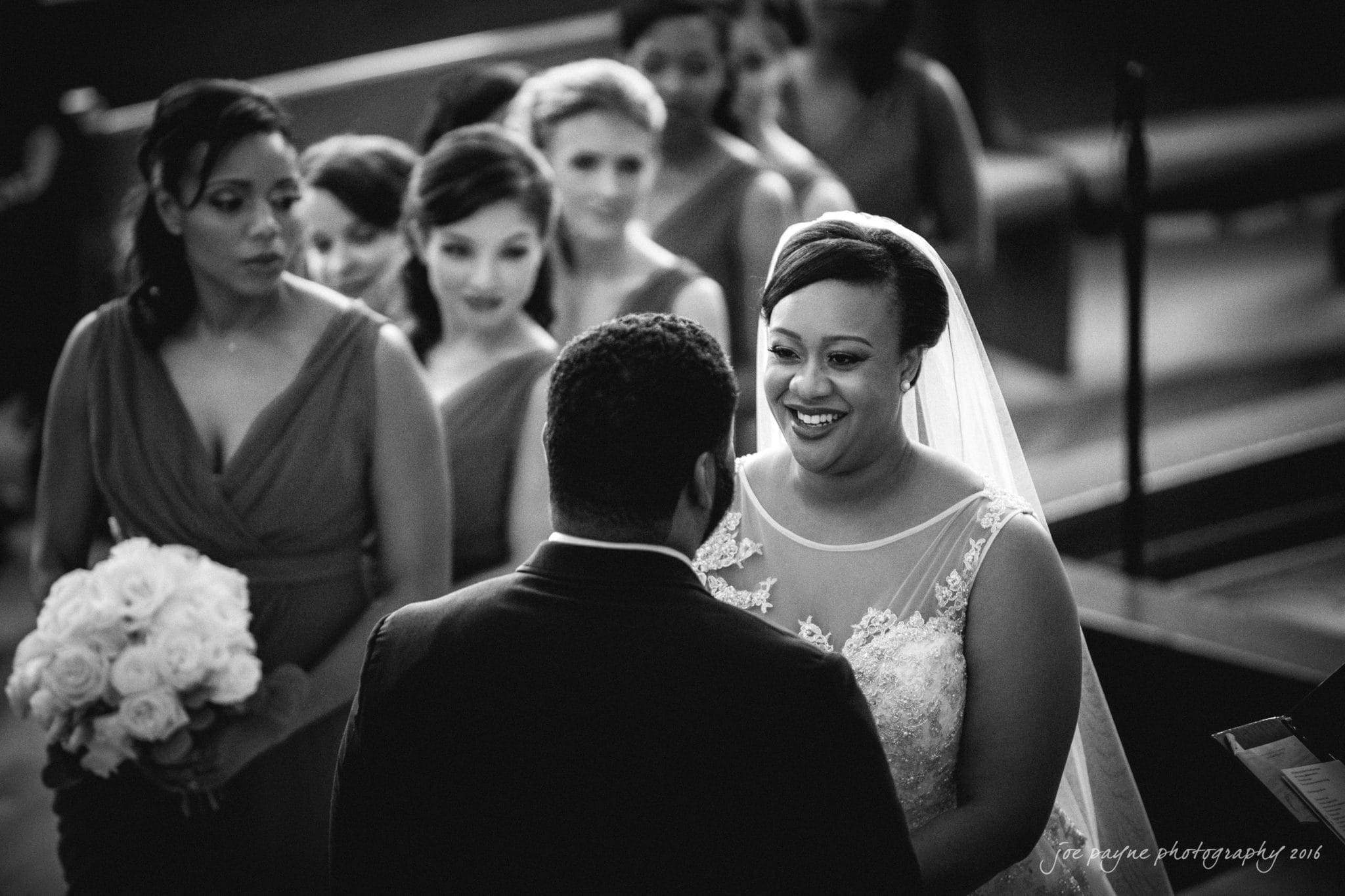 winston-salem nc wedding photography - toni & kevin A bride and groom stand facing each other, smiling during their wedding ceremony. Bridesmaids in matching dresses are in the background, holding bouquets. The setting appears to be a church or chapel.