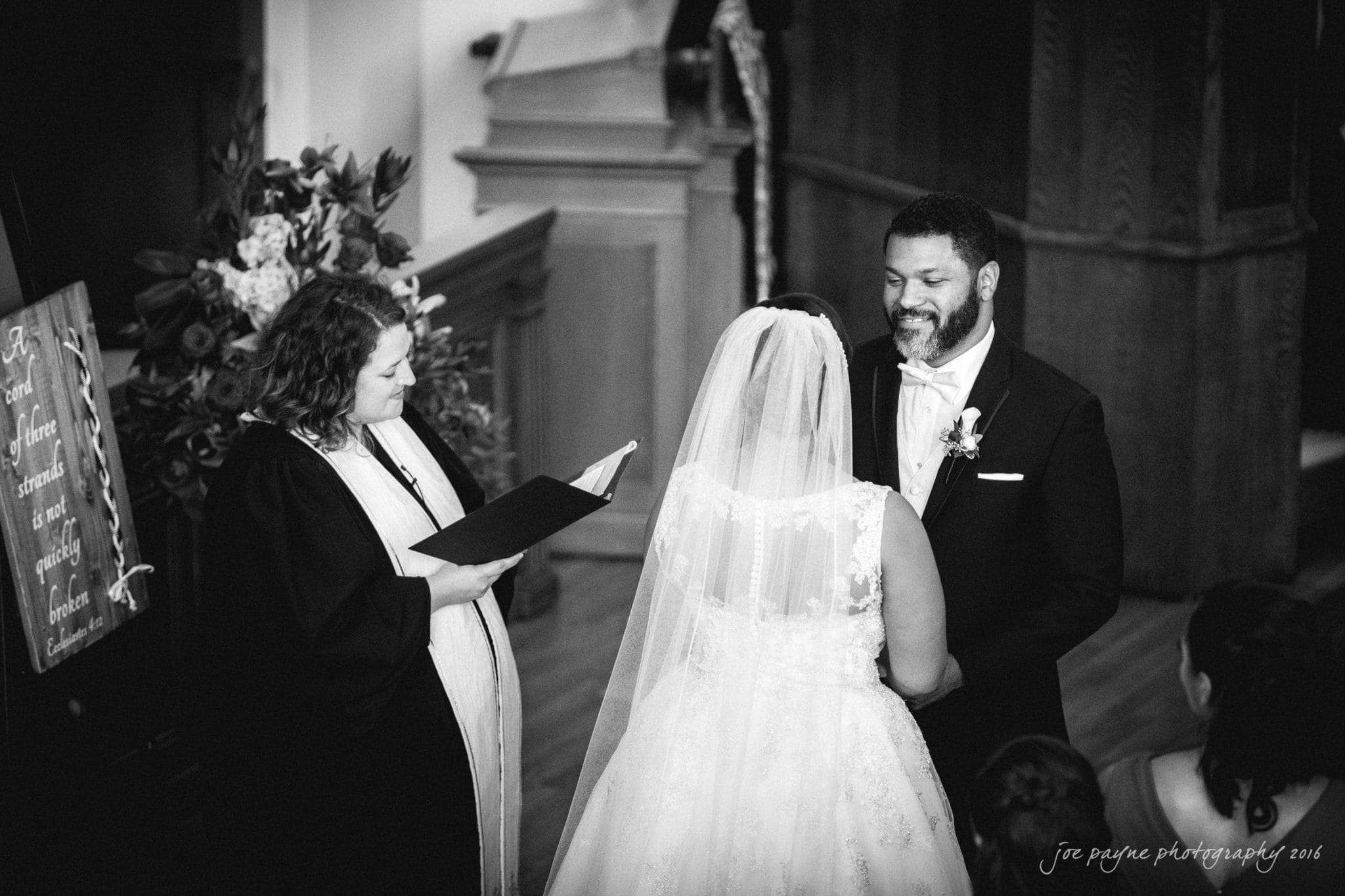 winston-salem nc wedding photography - toni & kevin A black-and-white photo of a wedding ceremony. A bride and groom stand facing each other, holding hands, with a wedding officiant reading from a book beside them. A floral arrangement and a decorative sign are visible in the background.