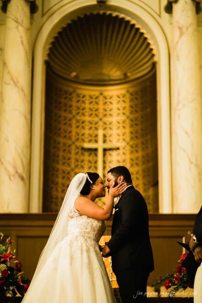 winston-salem nc wedding photography - toni & kevin A bride and groom stand at the altar in a church, exchanging vows. The bride is in a white gown with a veil, while the groom wears a black suit. A large cross and ornate archway are in the background, surrounded by floral arrangements.