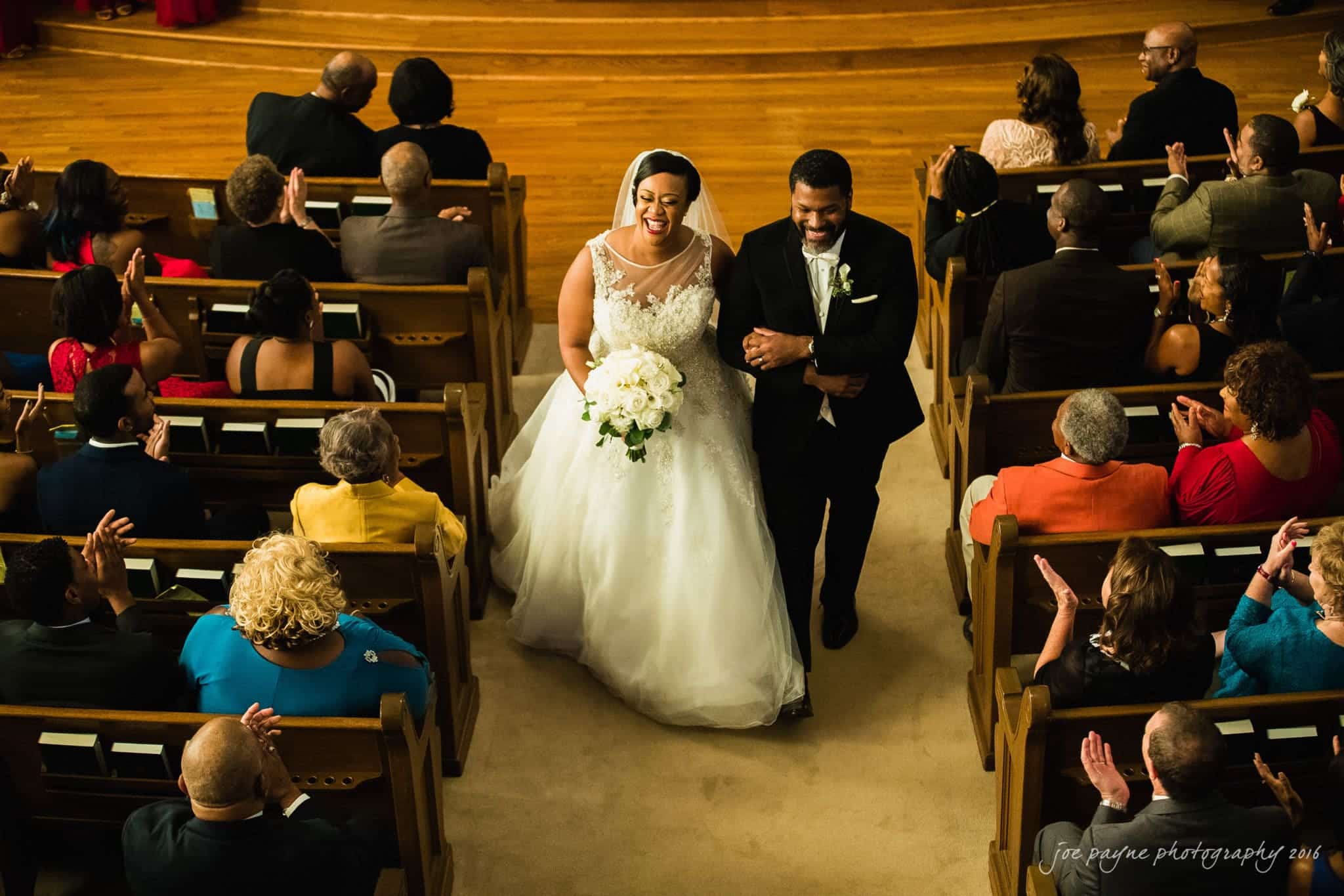 winston-salem nc wedding photography - toni & kevin A bride and groom smile and walk down the aisle, hand in hand, surrounded by applauding guests seated in wooden pews. The bride wears a white gown and veil, holding a bouquet, and the groom is in a black tuxedo.