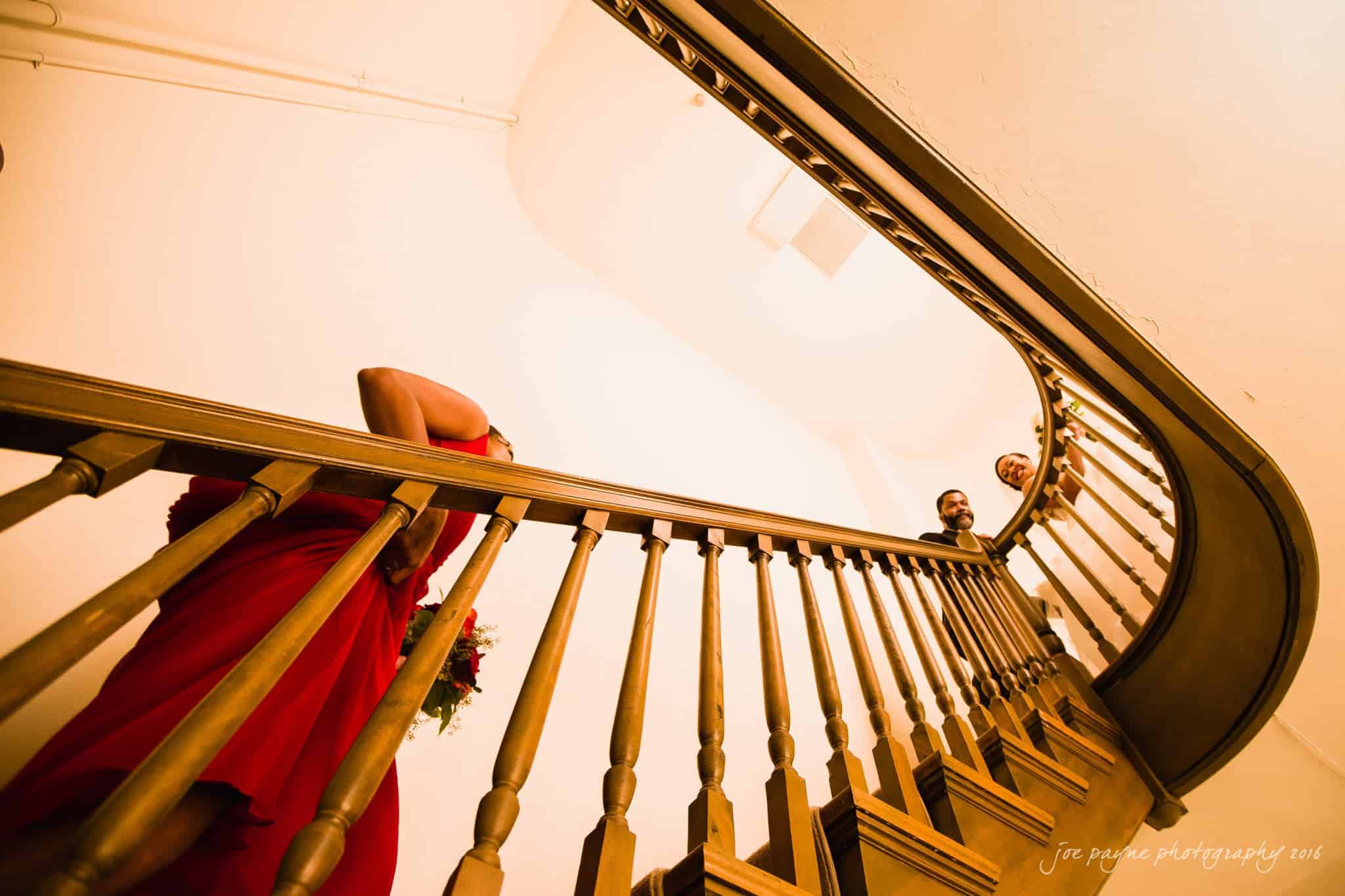 winston-salem nc wedding photography - toni & kevin A person in a red dress stands on a wooden staircase, holding a bouquet of flowers. Two people are visible above, looking down over the railing. The staircase curves gracefully upward, creating an elegant, sweeping perspective.