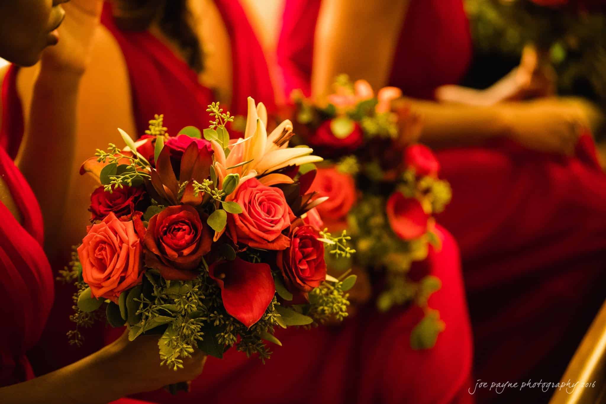 winston-salem nc wedding photography - toni & kevin Bridesmaids in red dresses hold vibrant bouquets of red and orange roses, lilies, and greenery. The close-up captures the rich colors and intricate details of the floral arrangements against the elegantly dressed attendants.