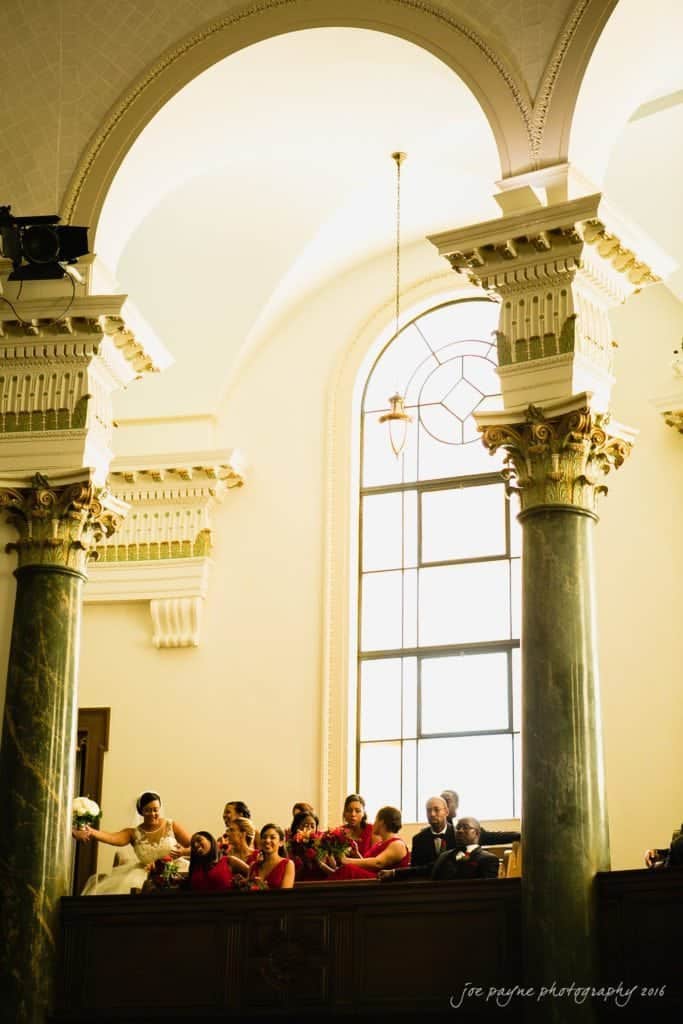 winston-salem nc wedding photography - toni & kevin Group of people in formal attire, with several women holding flowers, standing inside a building with large arched windows and ornate columns. The lighting is warm, creating an elegant atmosphere.