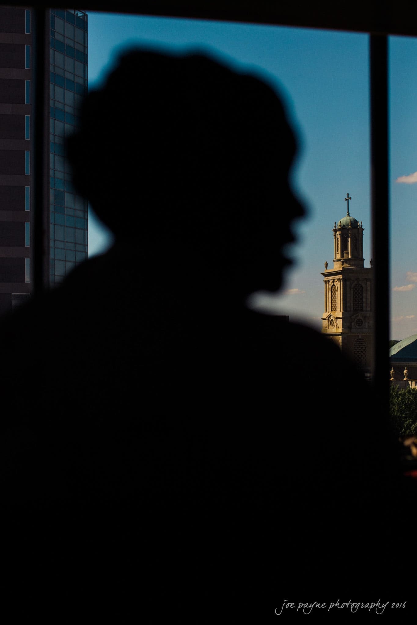 winston-salem nc wedding photography - toni & kevin Silhouette of a person looking out a window. In the background, a tall building and a church tower with a cross are visible against a blue sky.