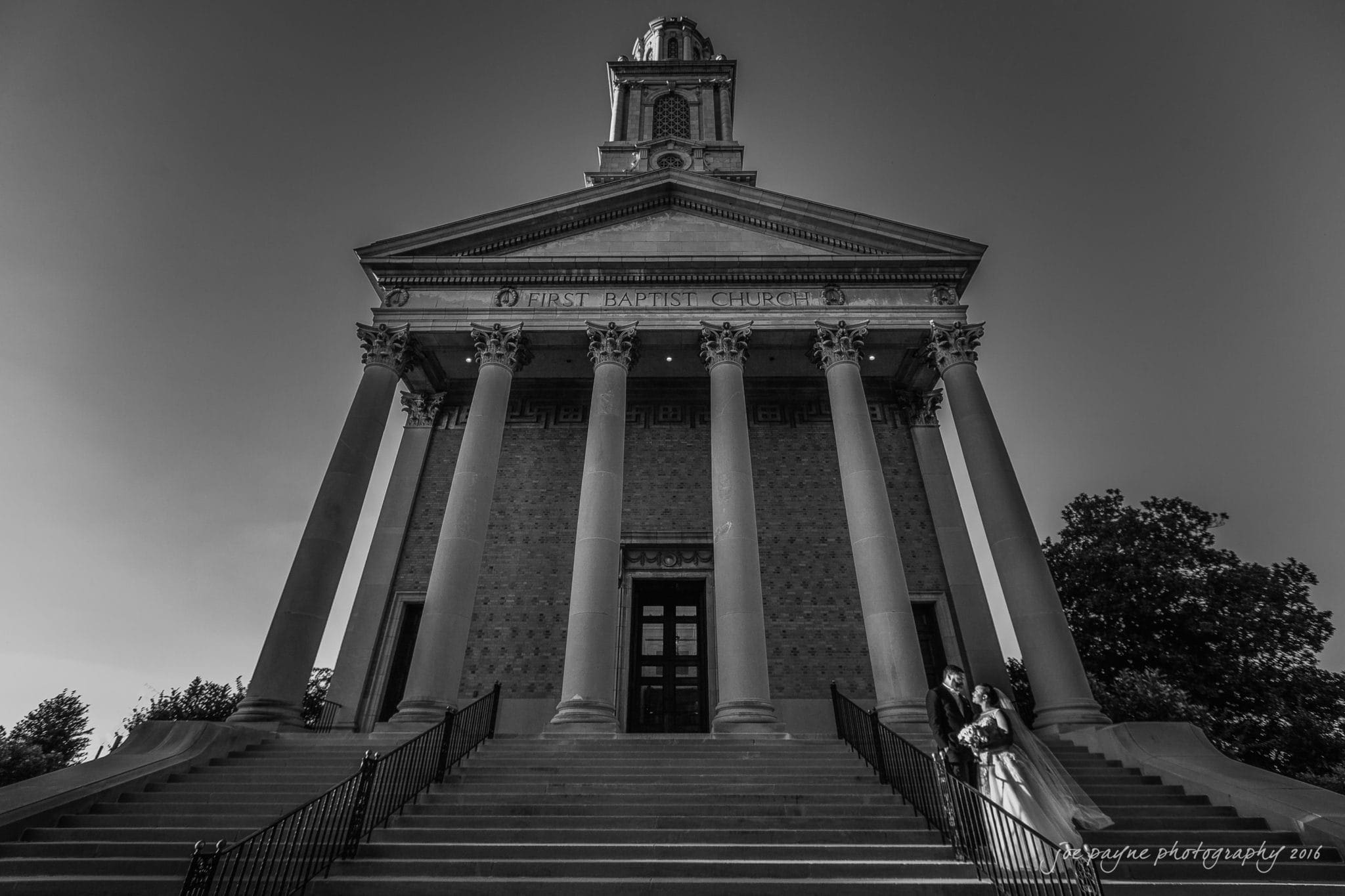 winston-salem nc wedding photography - toni & kevin A black and white image of a couple standing on the steps of a large building with tall columns. The sign reads First Baptist Church. They are dressed formally, and trees are visible in the background.