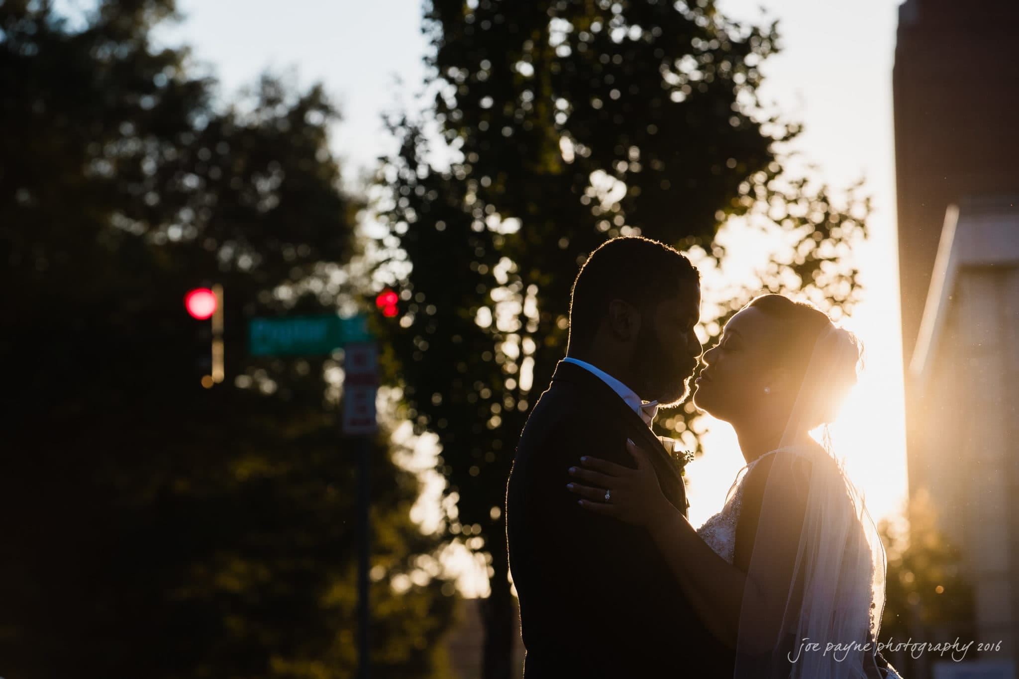 winston-salem nc wedding photography - toni & kevin Silhouetted couple in wedding attire sharing a tender moment outdoors at sunset. Trees and a street sign are visible in the background, with a soft golden light illuminating their outlines.
