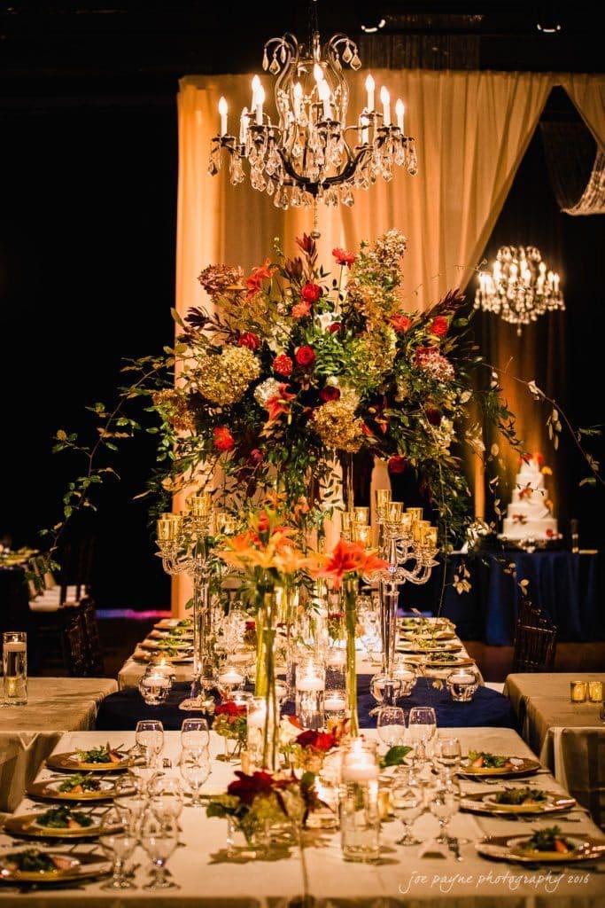 winston-salem nc wedding photography - toni & kevin An elegant banquet table is adorned with lush floral centerpieces, candles, and fine dining settings. A grand chandelier hangs overhead, casting a warm glow. The background features draped fabric and another chandelier, adding to the opulent ambiance.