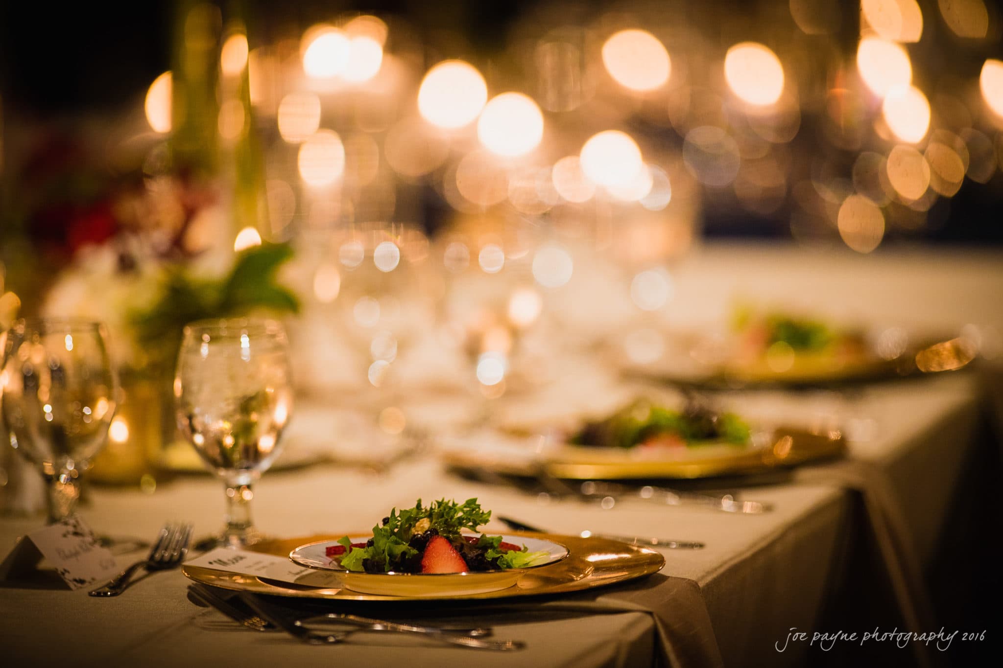 winston-salem nc wedding photography - toni & kevin A beautifully set dining table with a blurred bokeh background. The table features elegant glassware and plates with fresh salads. Warm, glowing lights create a romantic atmosphere.