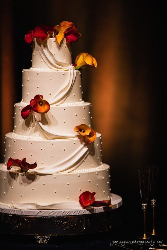 winston-salem nc wedding photography - toni & kevin A five-tiered white wedding cake adorned with orange and red flowers. The cake is embellished with draped fondant and small pearl-like accents. Two champagne flutes are nearby, and the background is softly lit.