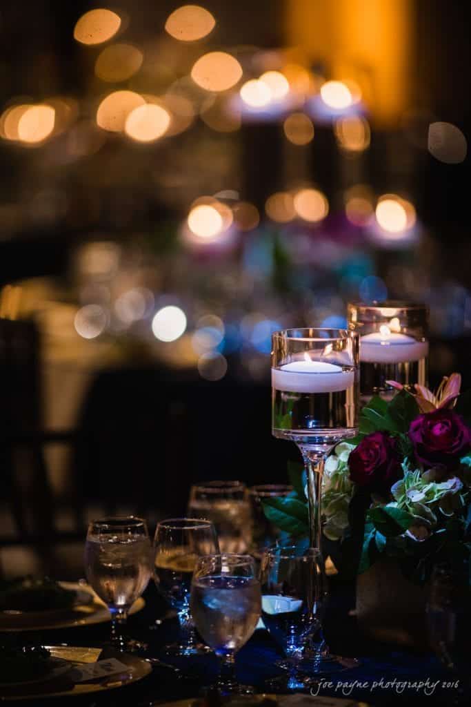 winston-salem nc wedding photography - toni & kevin A dimly lit, elegant dinner setting with floating candles in tall holders, reflecting soft, warm bokeh lights. The table features glassware, plates, and a floral centerpiece with red and purple flowers.