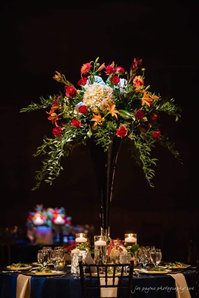 winston-salem nc wedding photography - toni & kevin A tall, elegant floral centerpiece with red roses, orange lilies, greenery, and white hydrangeas in a dark vase on a table set with candles, glassware, and plates. A dimly lit, sophisticated background adds to the ambiance.